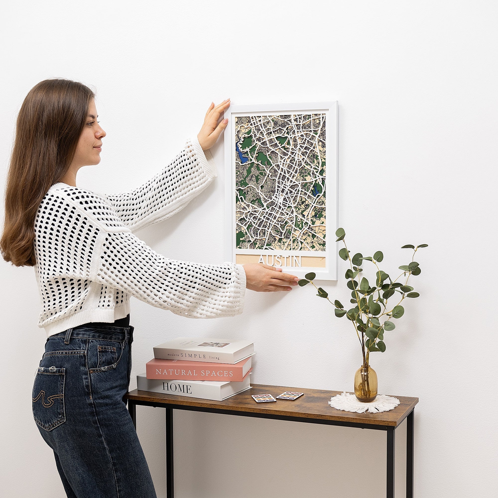 Woman hanging a framed map on a white wall with a small table and plant in the foreground.
