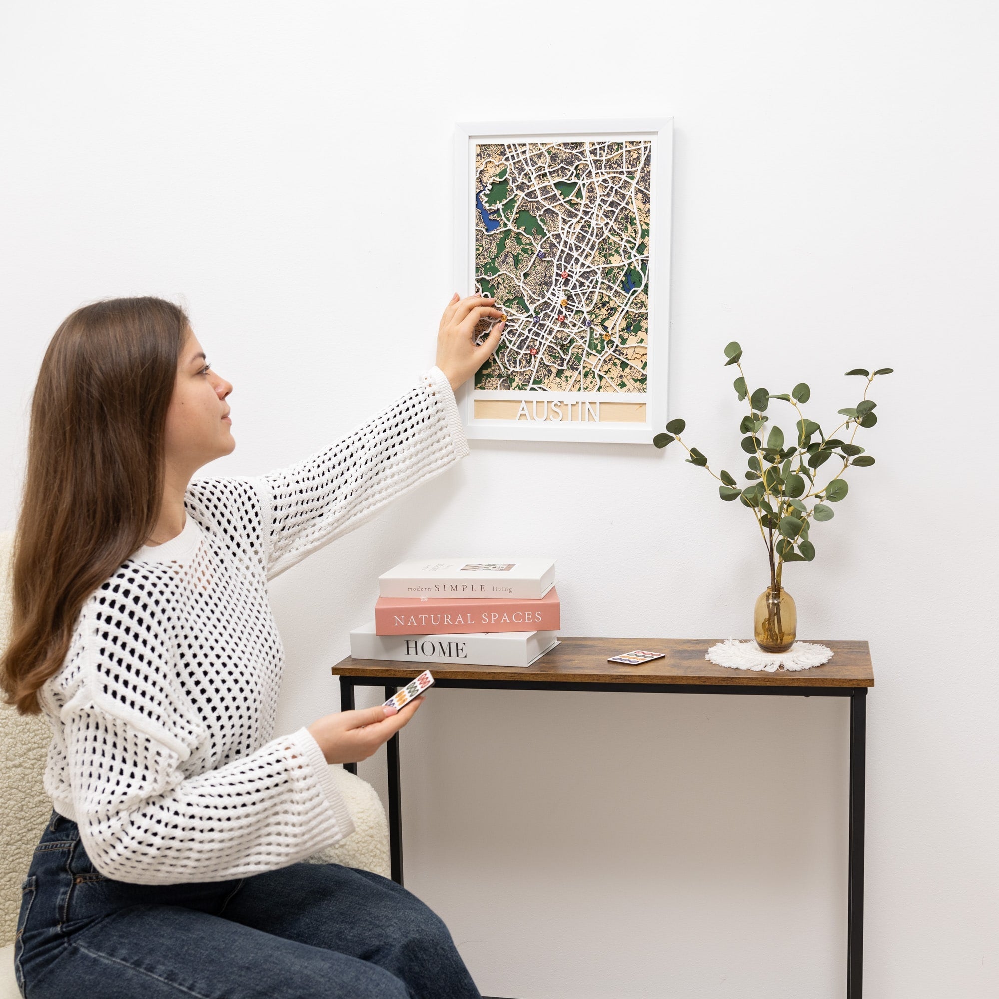 Woman hanging a framed map on a wall above a small table with books and a plant.