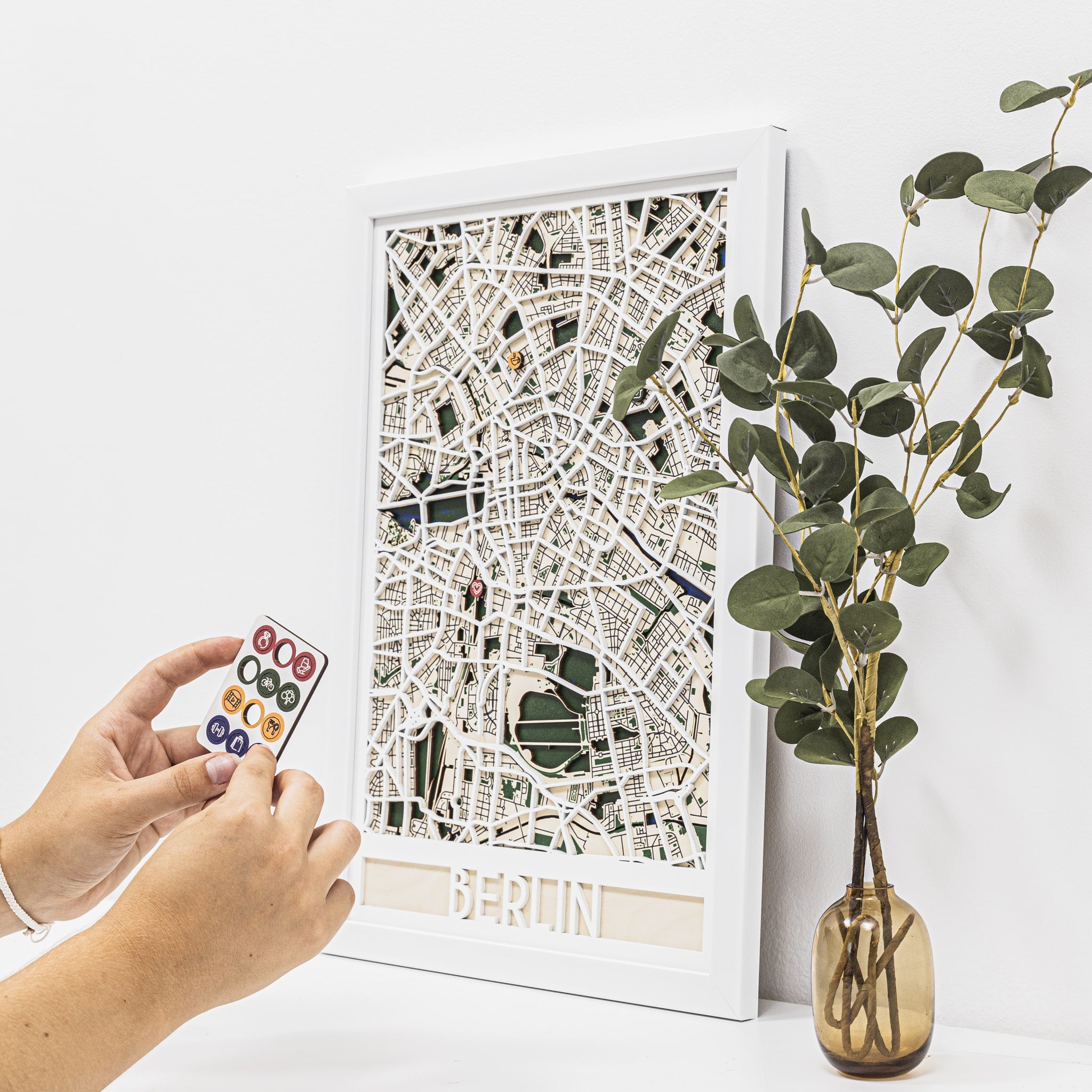 Framed city map of Berlin with a hand holding a small colorful object and a vase with greenery on a white background.