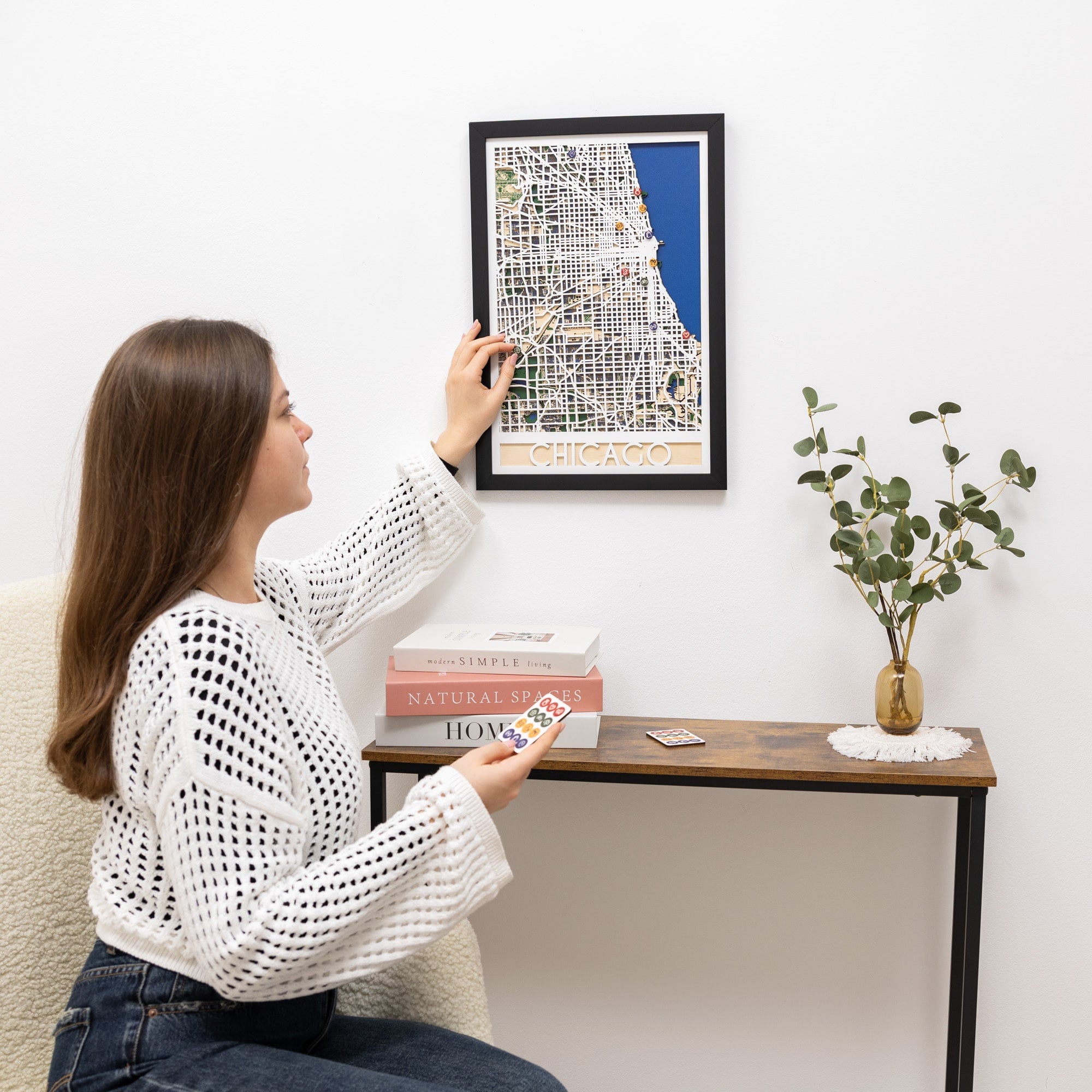 Woman hanging a framed map on a wall above a table with books and a plant.
