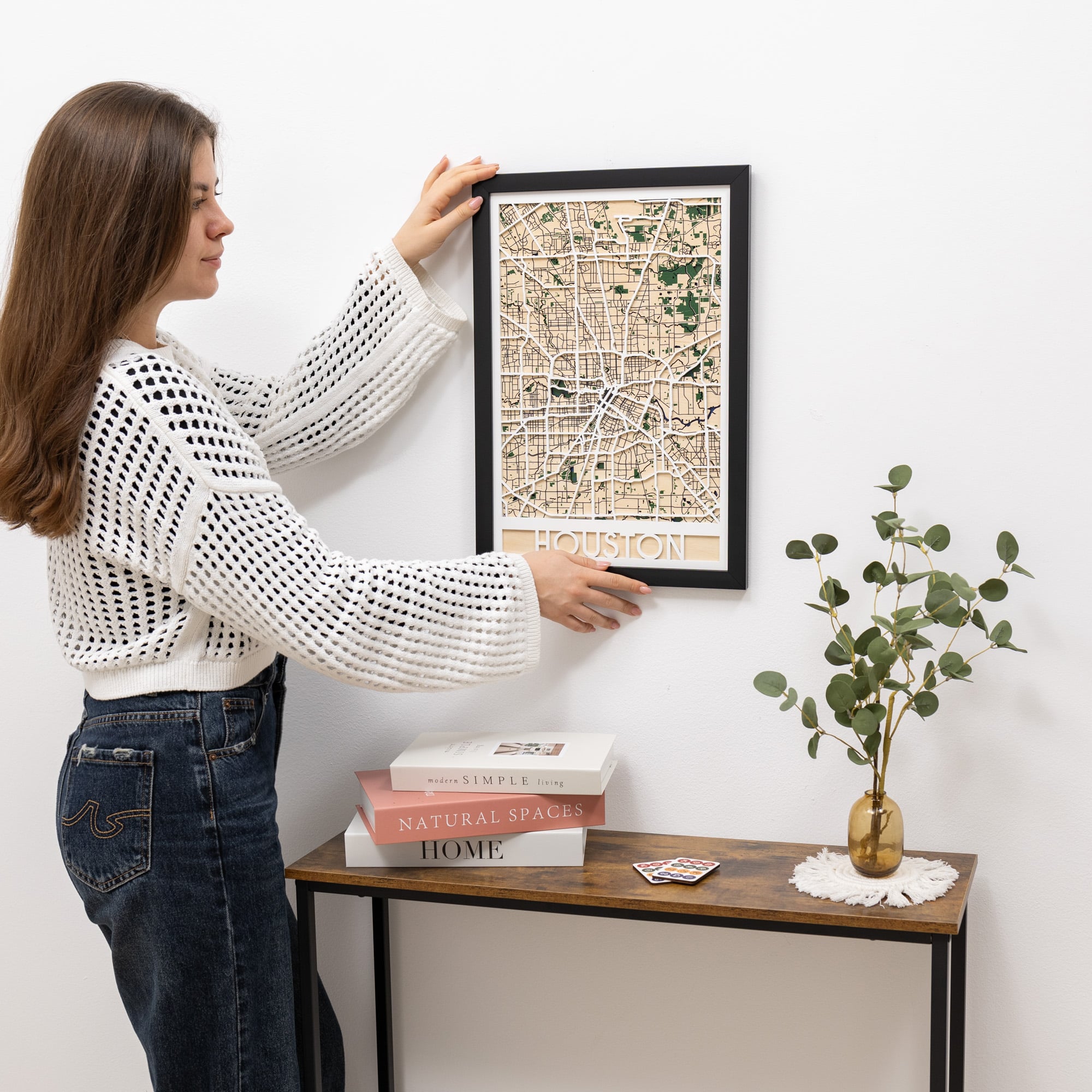 Woman hanging a framed map of Houston on a wall next to a small table with books and a plant.