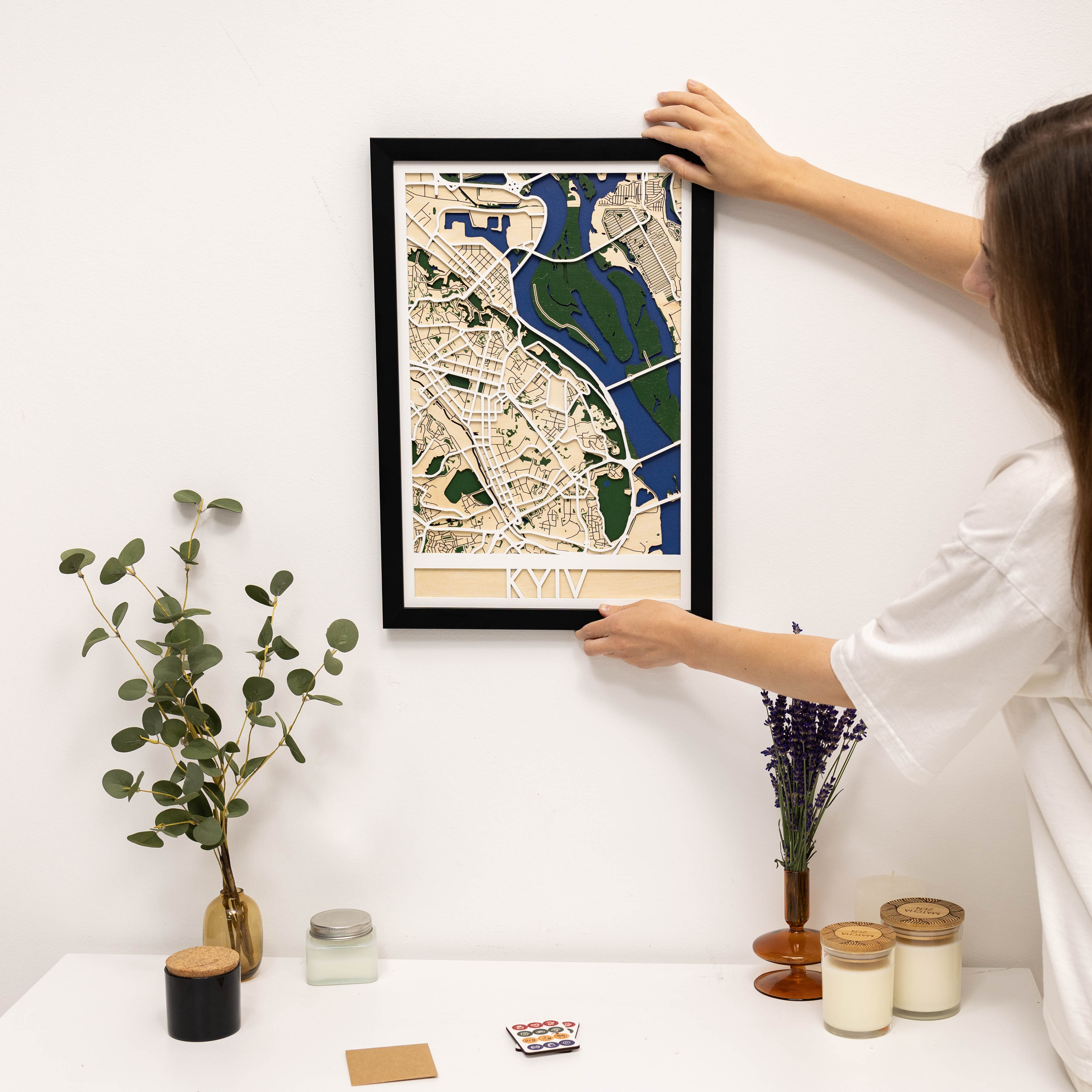 Person hanging a framed map of Kyiv on a wall with decorative items below.