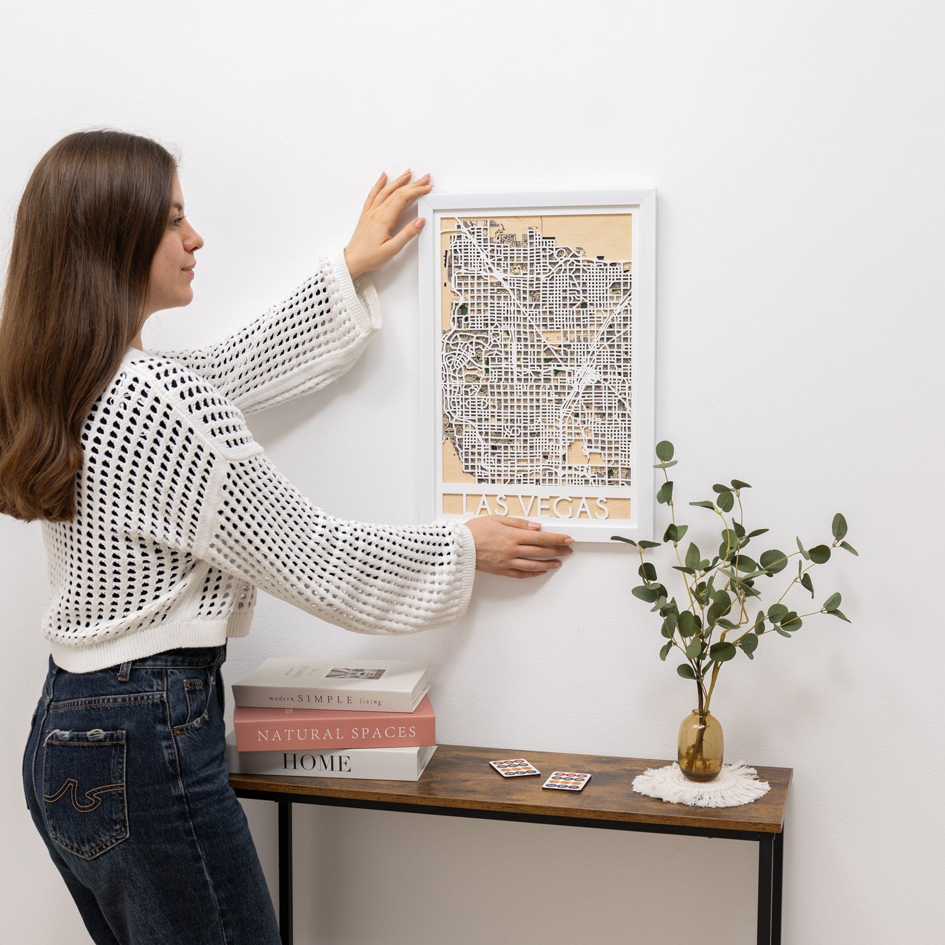 Woman hanging a framed map on a wall next to a wooden table with books and a plant.