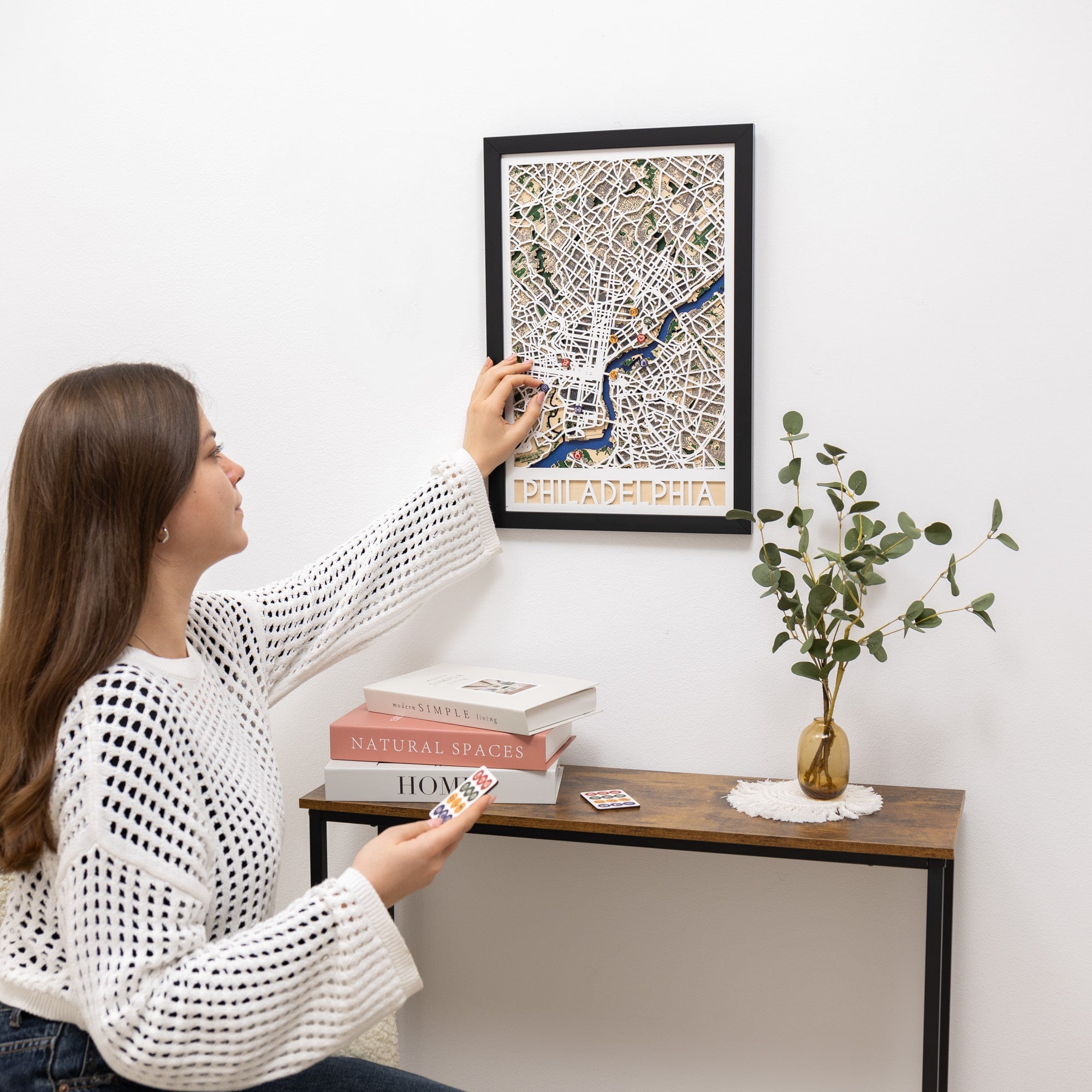 Woman hanging a framed map of Philadelphia on a wall above a small table with books and a plant.