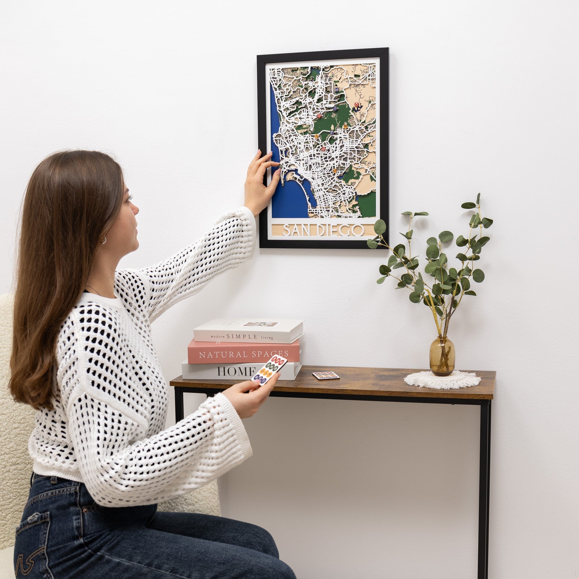 Woman hanging a framed map on a wall above a small table with decor items.