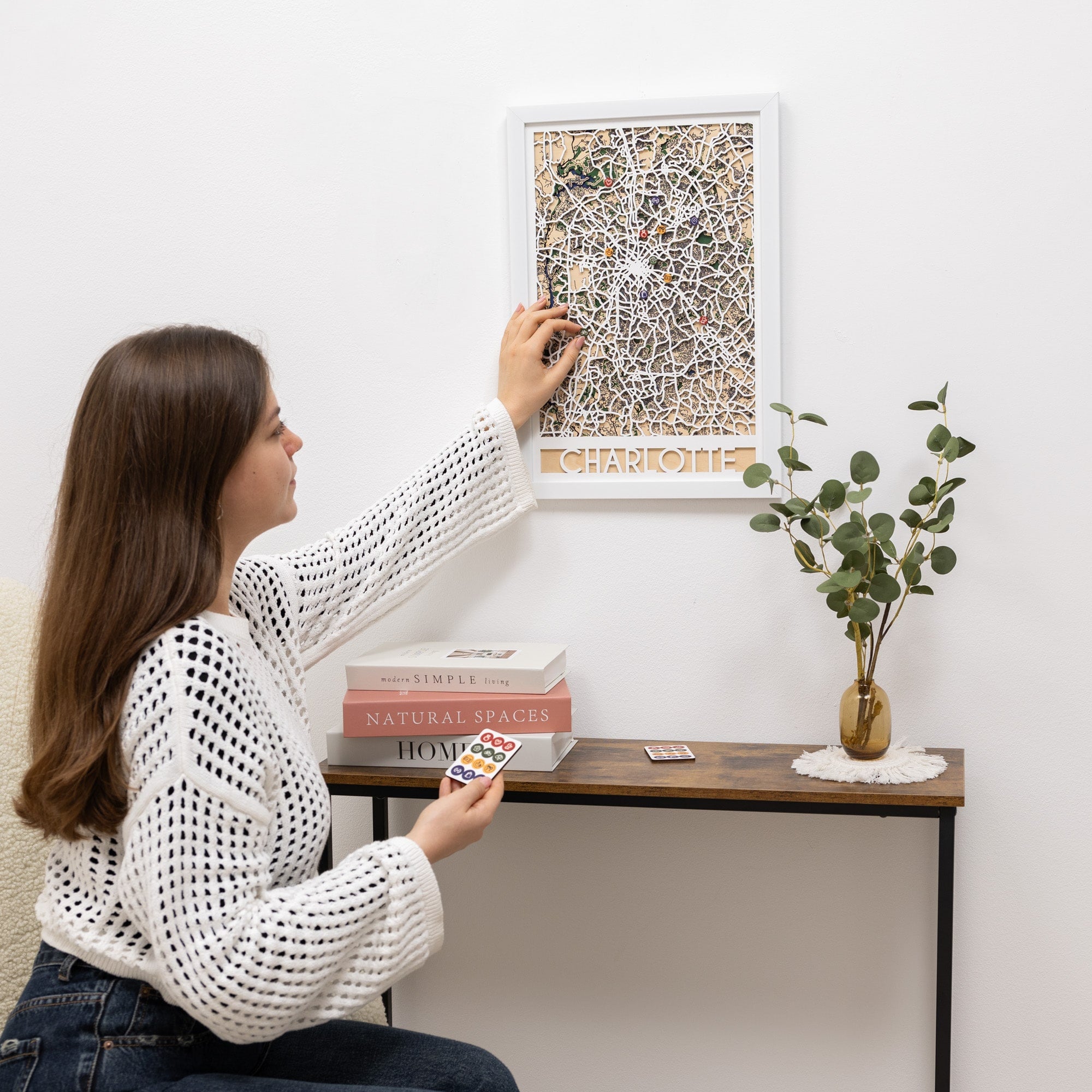 Woman hanging a framed artwork on a wall next to a small table with decor items.