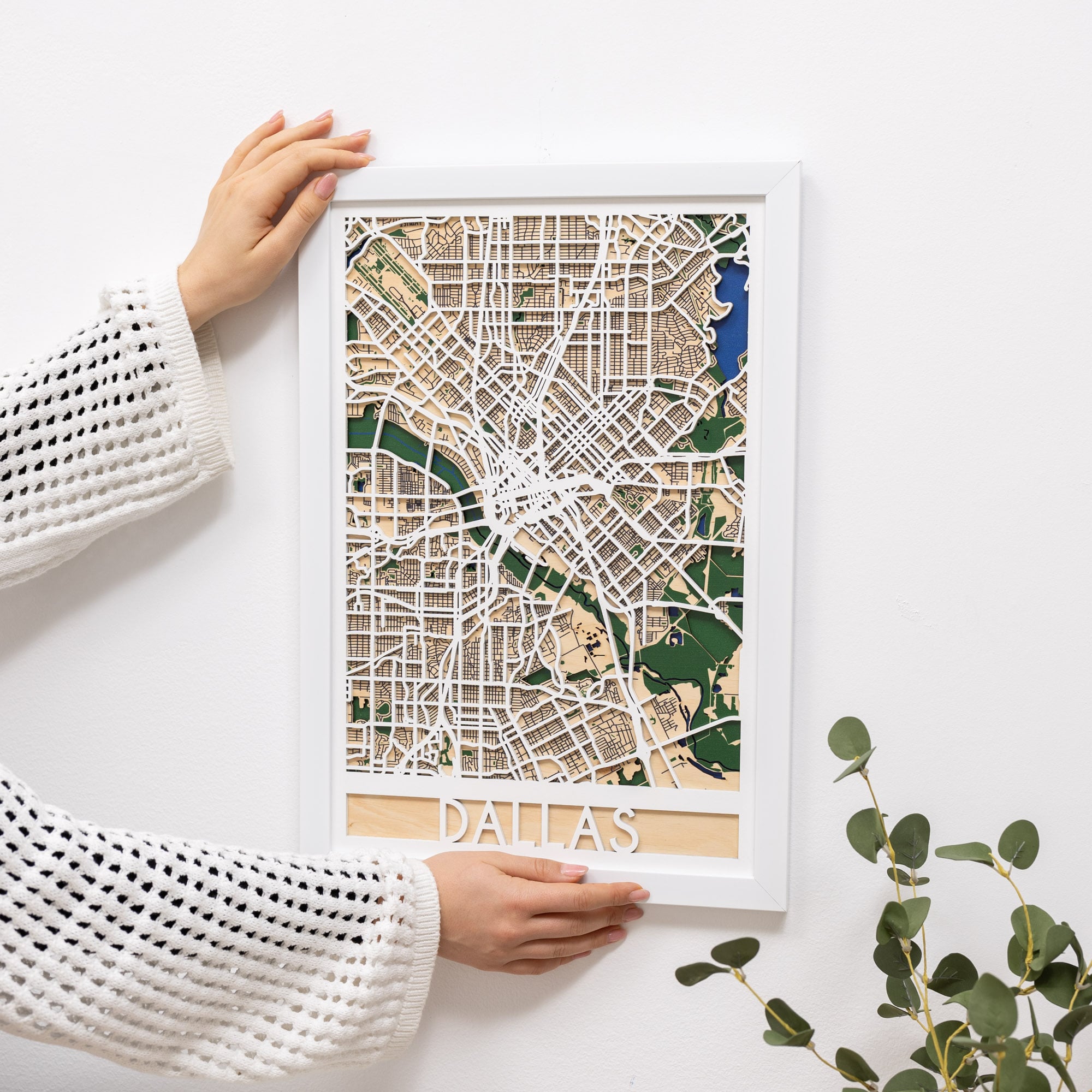 Woman hanging a framed map on a wall next to a wooden side table with books and a plant.
