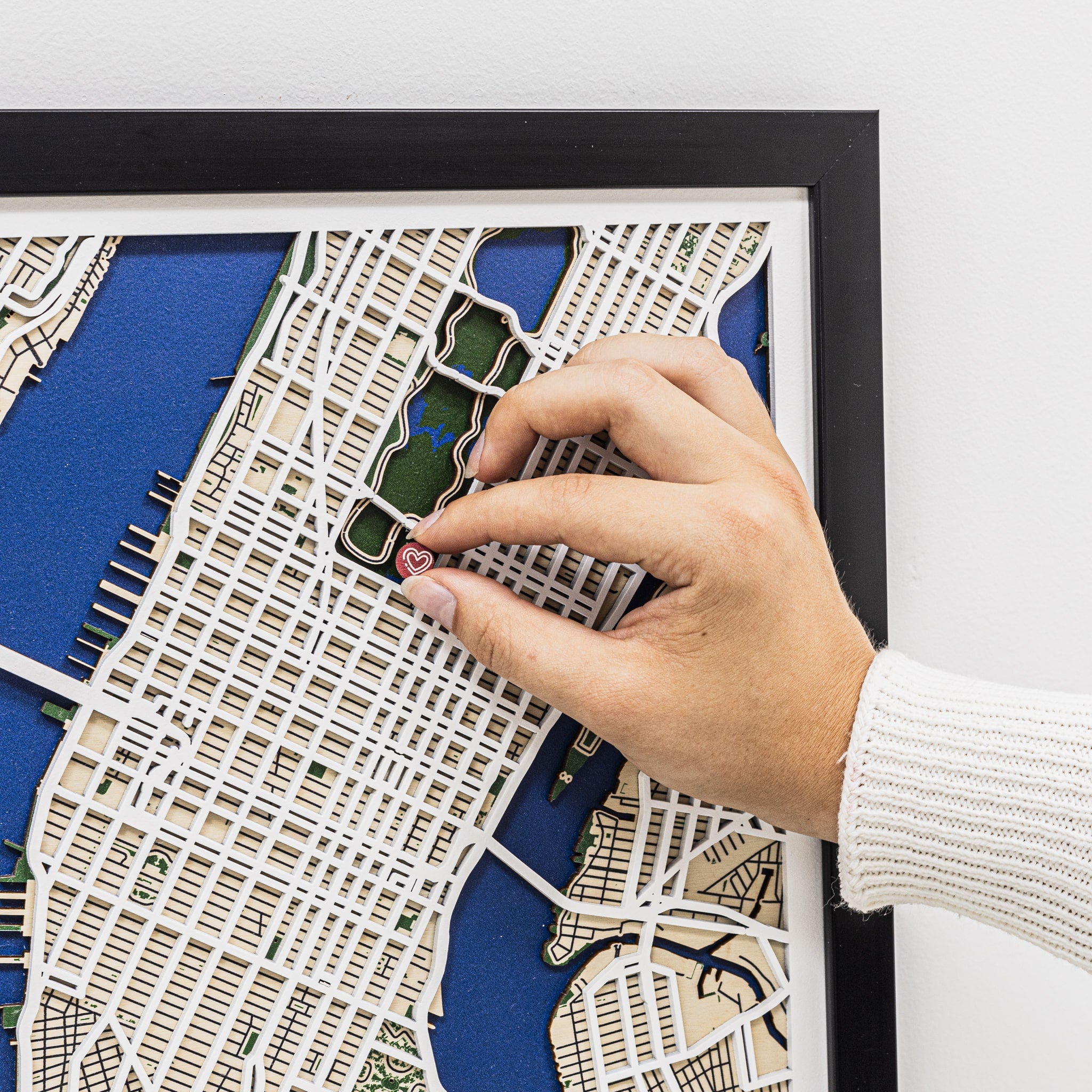 Hand placing a heart-shaped pin on a detailed map in a black frame.