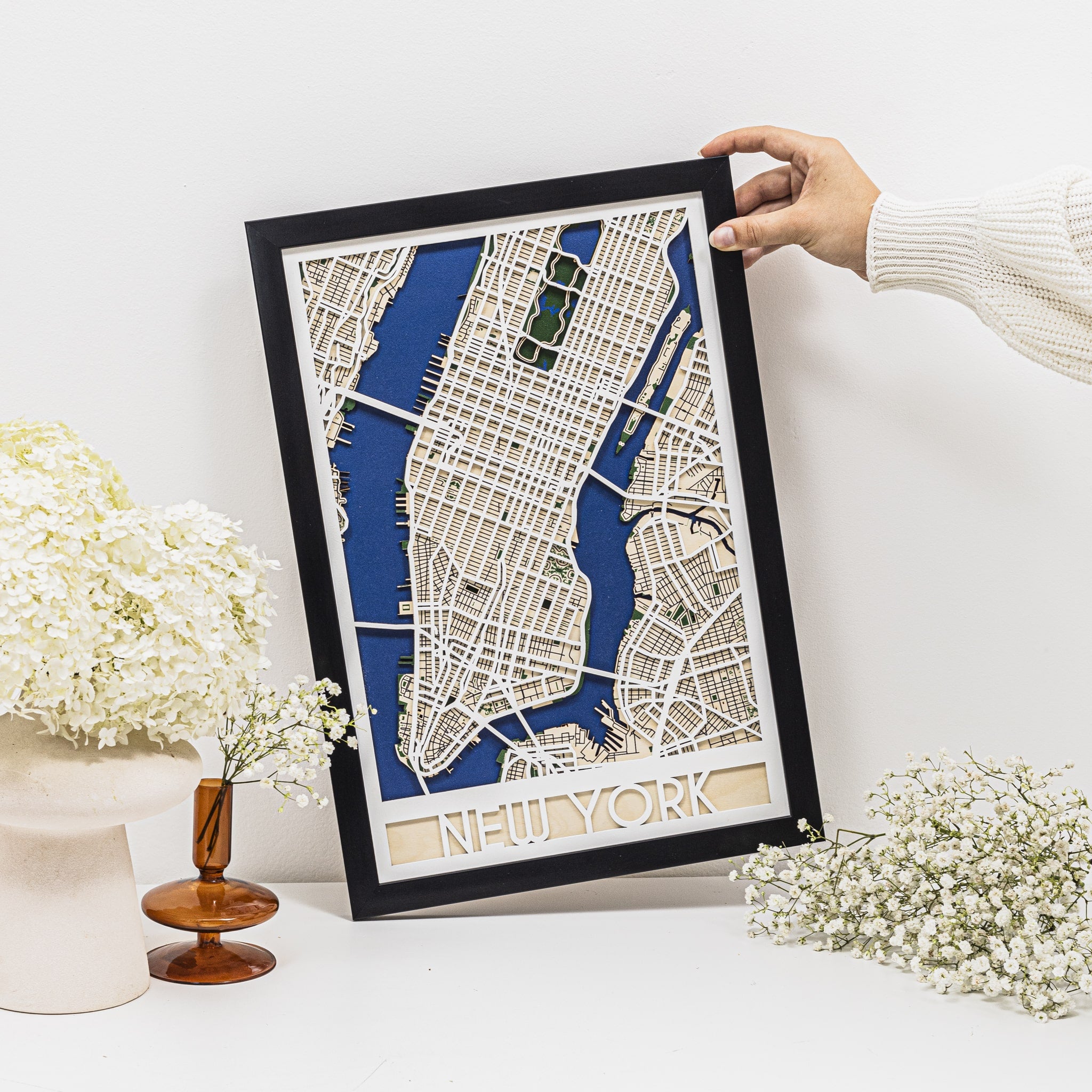 Framed New York city map held by a person with decorative elements on a white surface.