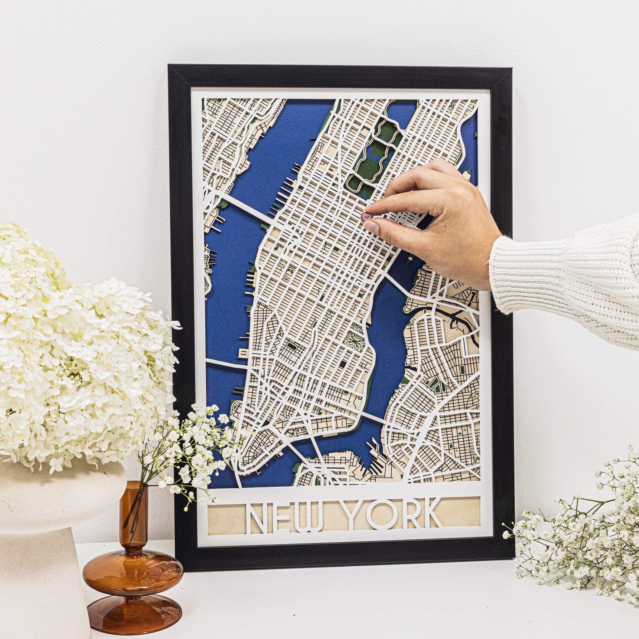 Framed map of New York with a hand pointing at it on a white surface with flowers.