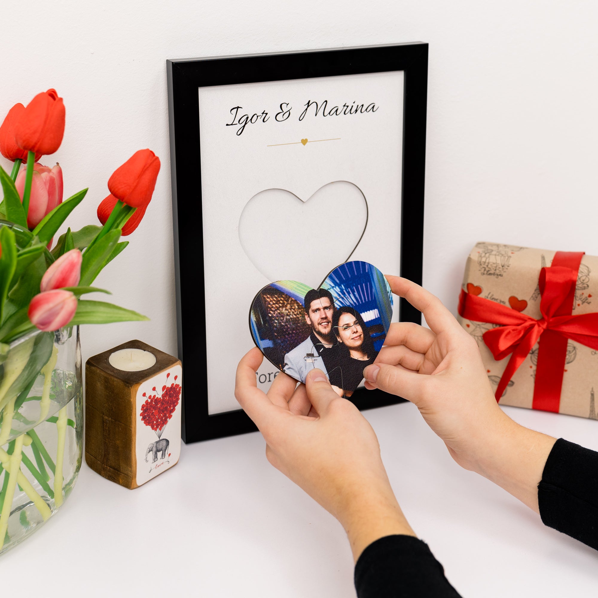 Person holding a heart-shaped photo cutout with a framed picture of two people, surrounded by red tulips and a gift box.
