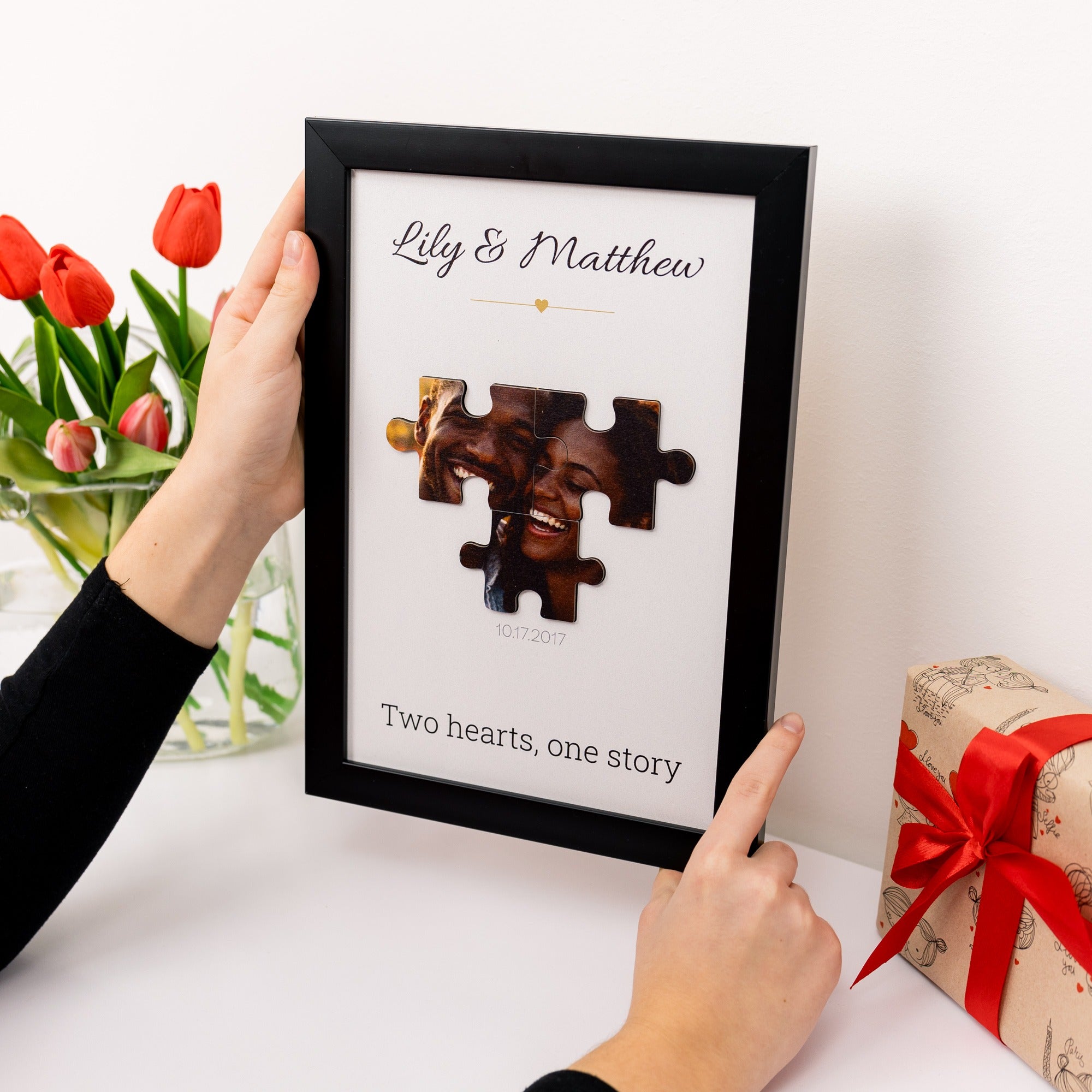 Framed photo of two puzzle pieces with a couple's names and a gift box with a red ribbon on a white surface.