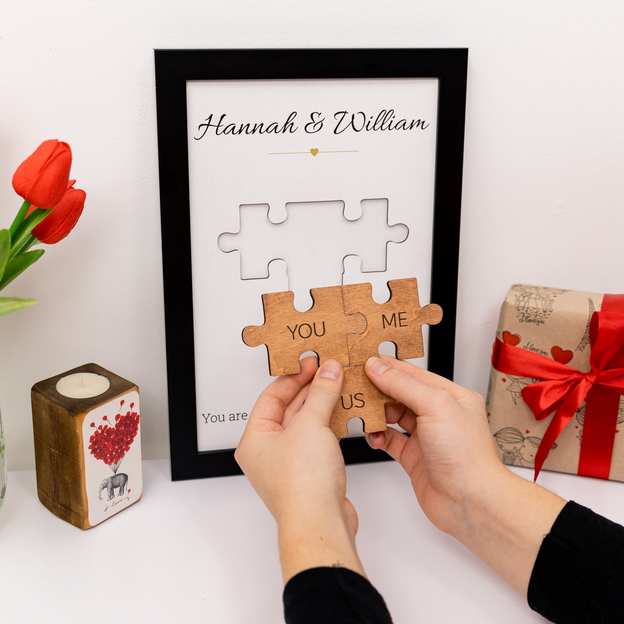 Wooden puzzle piece with 'You & Me = Us' text, framed with 'Hannah & William', on a white surface with decorative items.