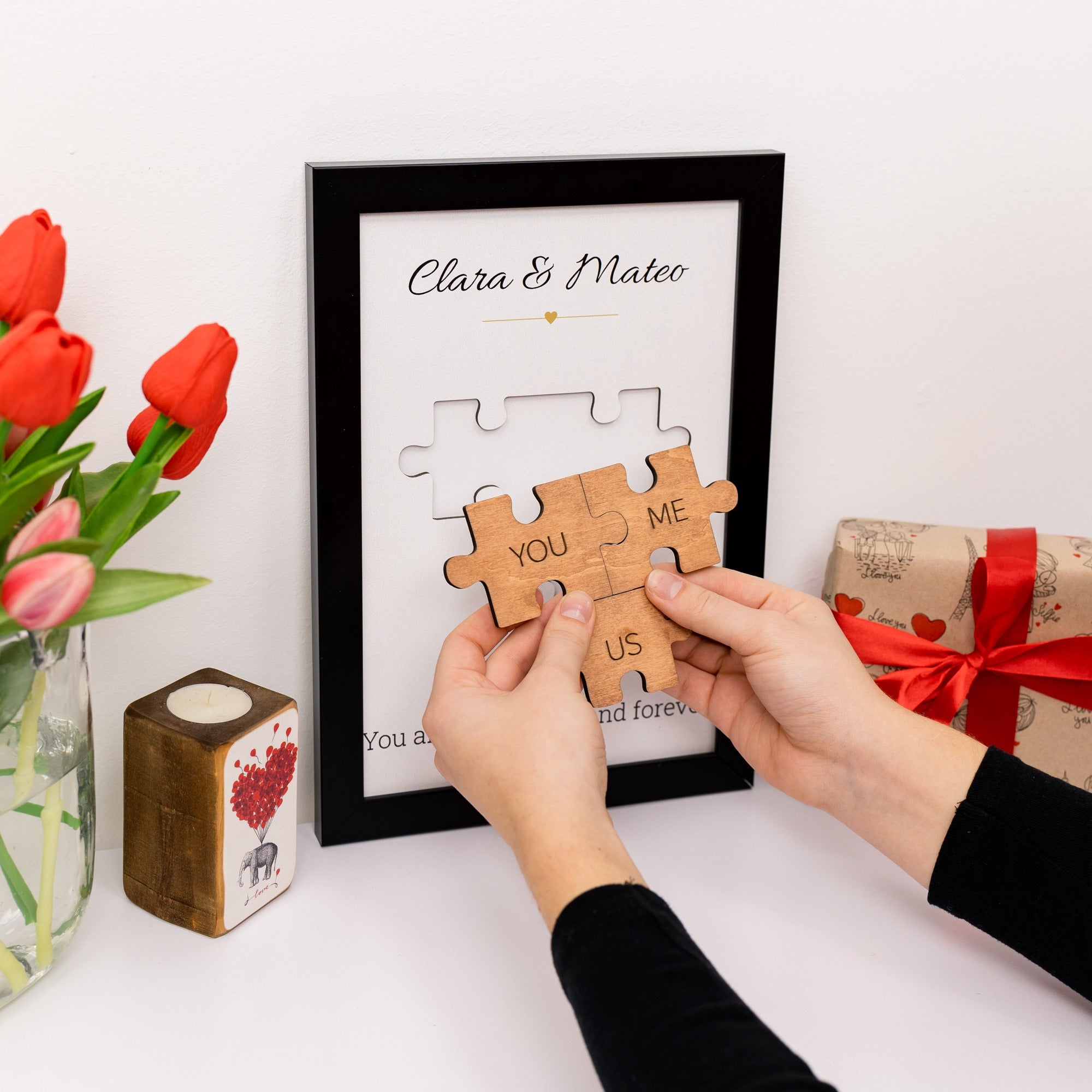 Person assembling a wooden puzzle piece with text in a framed picture, surrounded by red tulips and gift box.