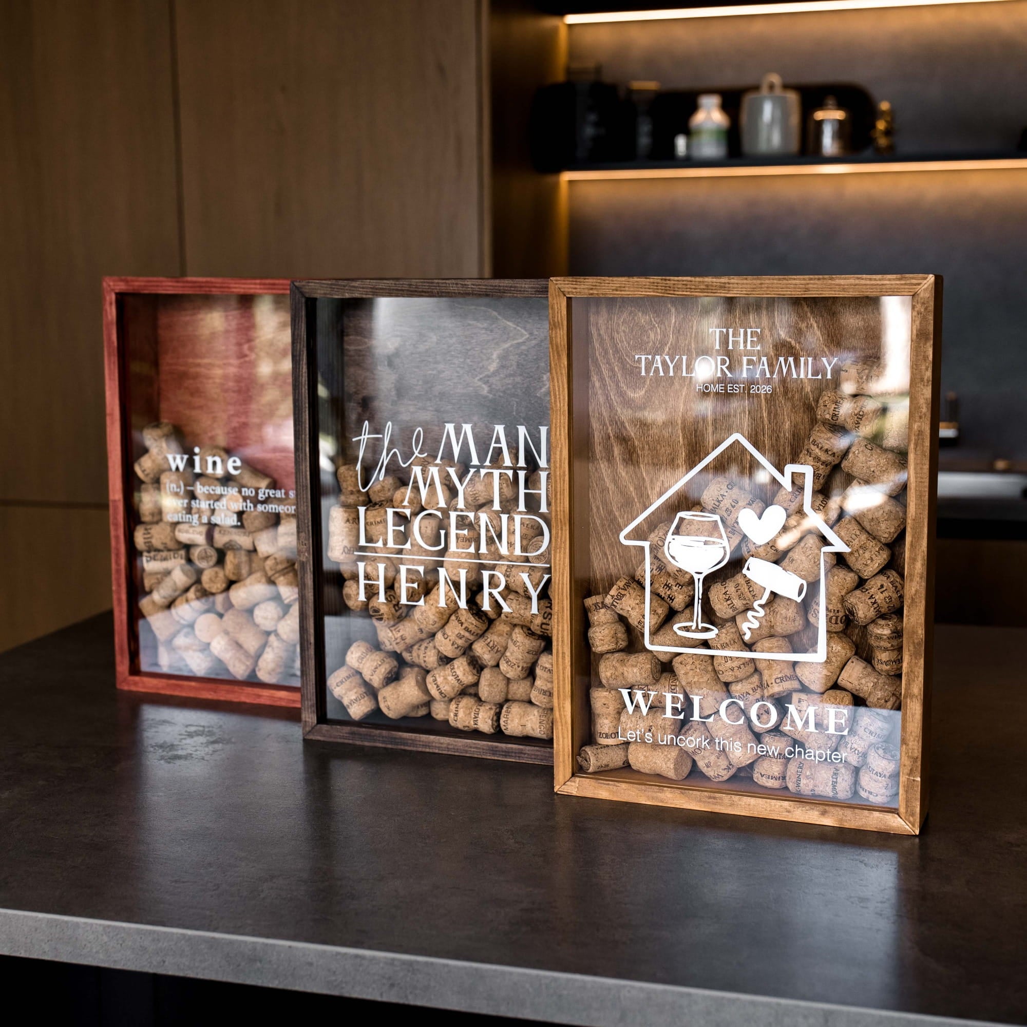 Three personalized framed wine cork collections displayed on a dark countertop in a kitchen setting.