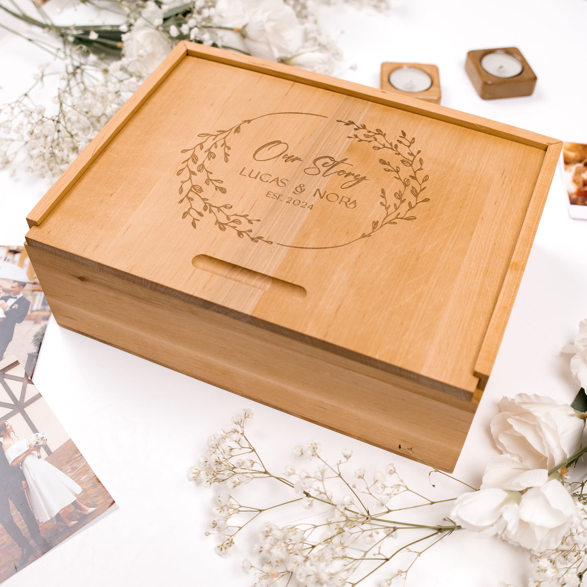 Wooden box with engraved text 'Our Story Lucas & Nora DE 2024' on a white surface with flowers and photos.