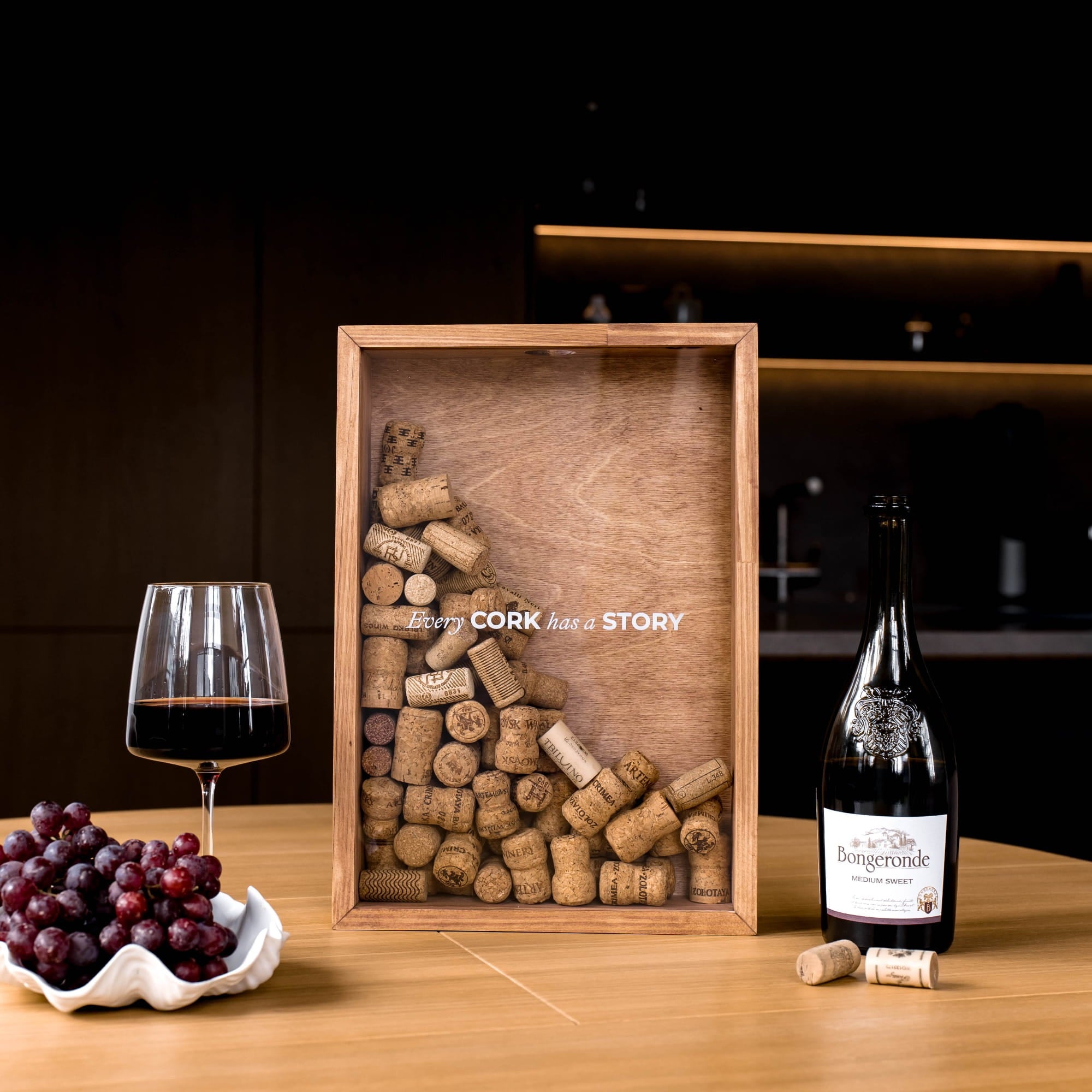 A wooden framed display filled with wine corks sits on a tabletop beside a glass of red wine, a bowl of grapes, and a wine bottle. 