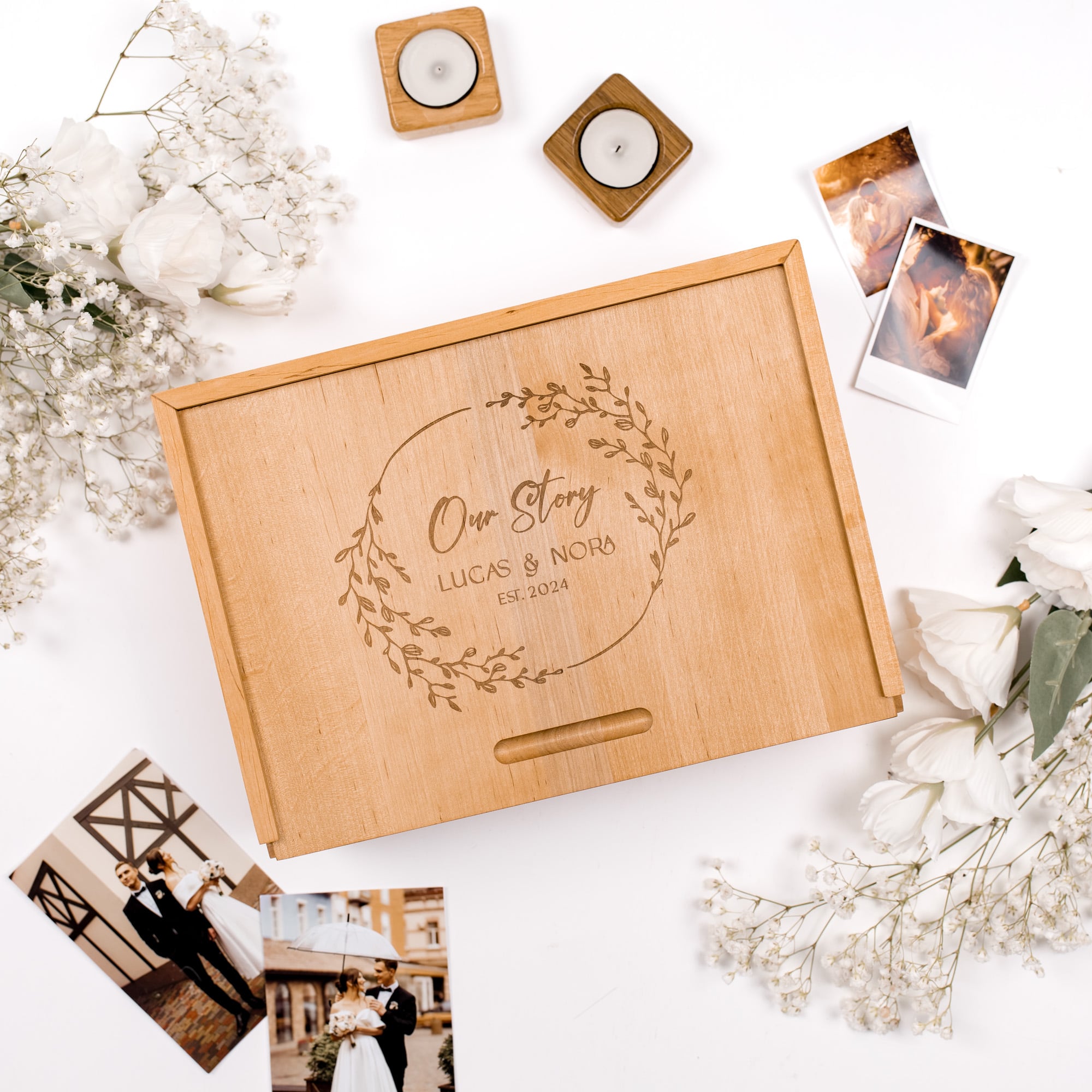 Wooden box with engraved text 'Our Story Lucas & Nora' on a white surface with flowers and photos.