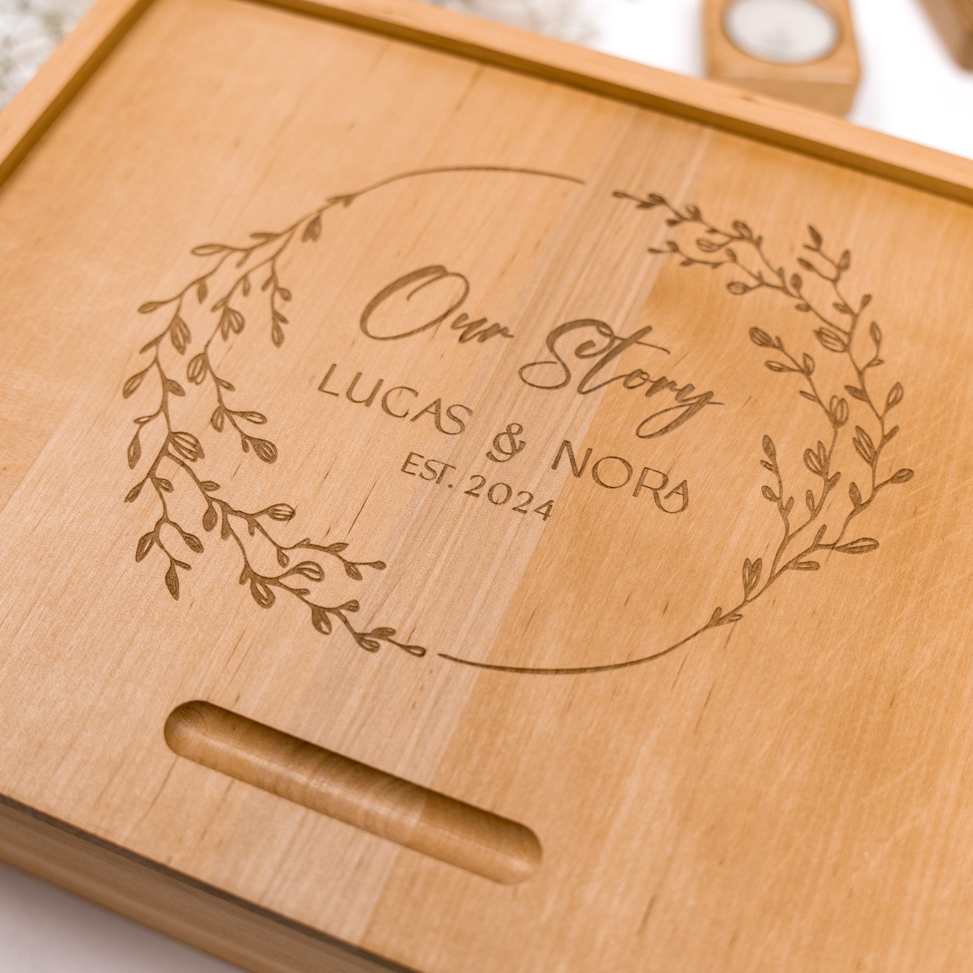 Wooden cutting board with engraved text 'Our Story LUCAS & NORA EST. 2024' on a light background.