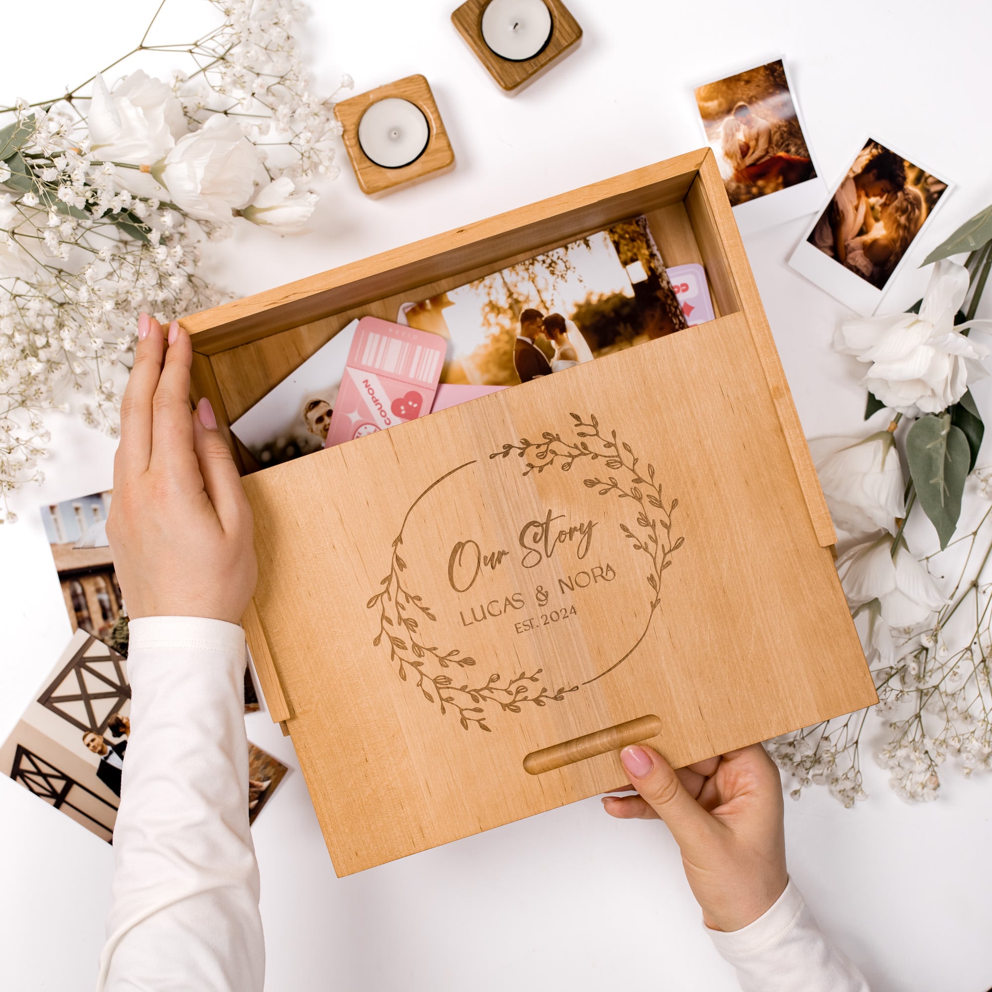 Wooden photo box with 'Our Story' engraving, held by a person, surrounded by photos and flowers on a white background.
