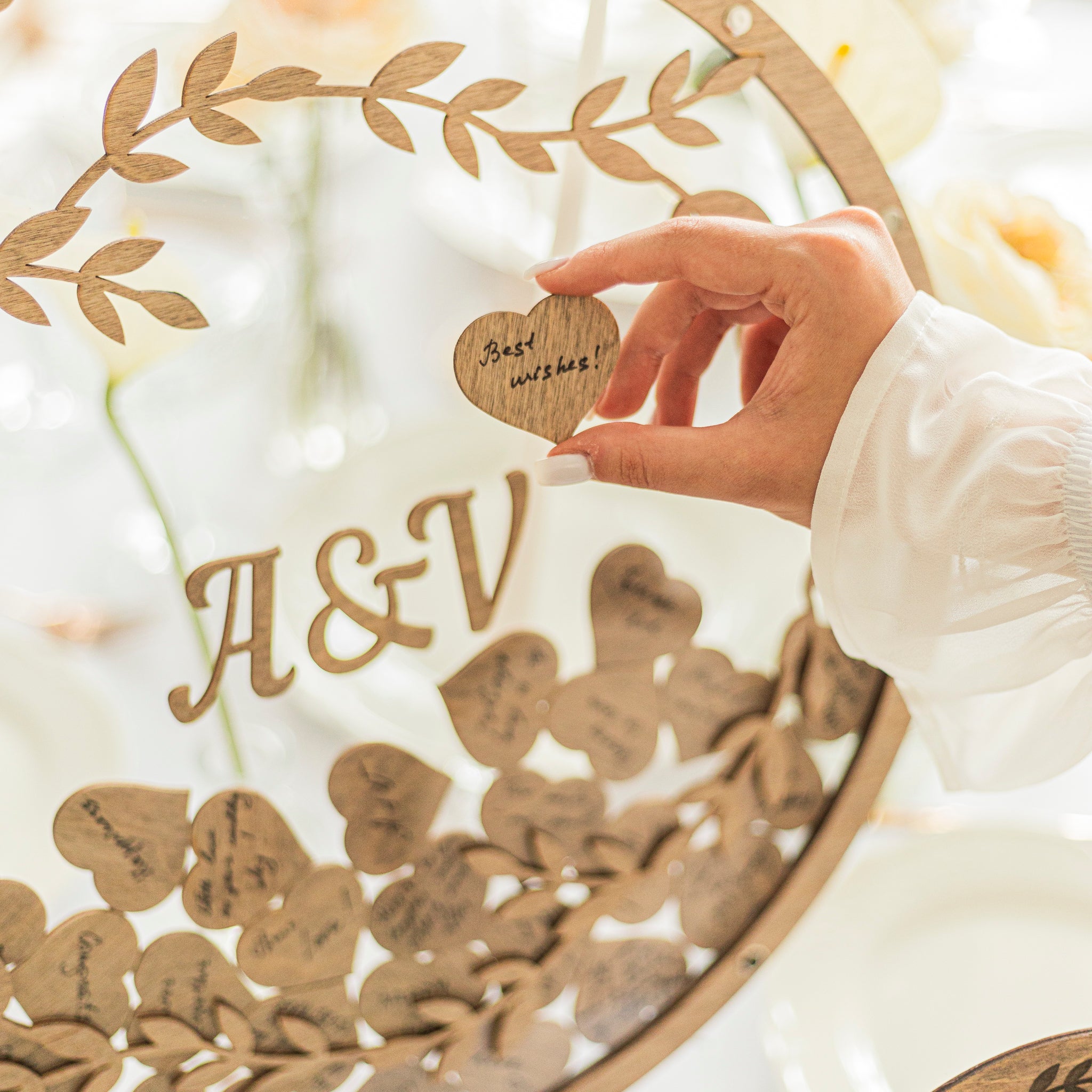 Hand holding a heart-shaped wooden tag with 'Best wishes!' over a decorative wreath with 'A & V' initials.