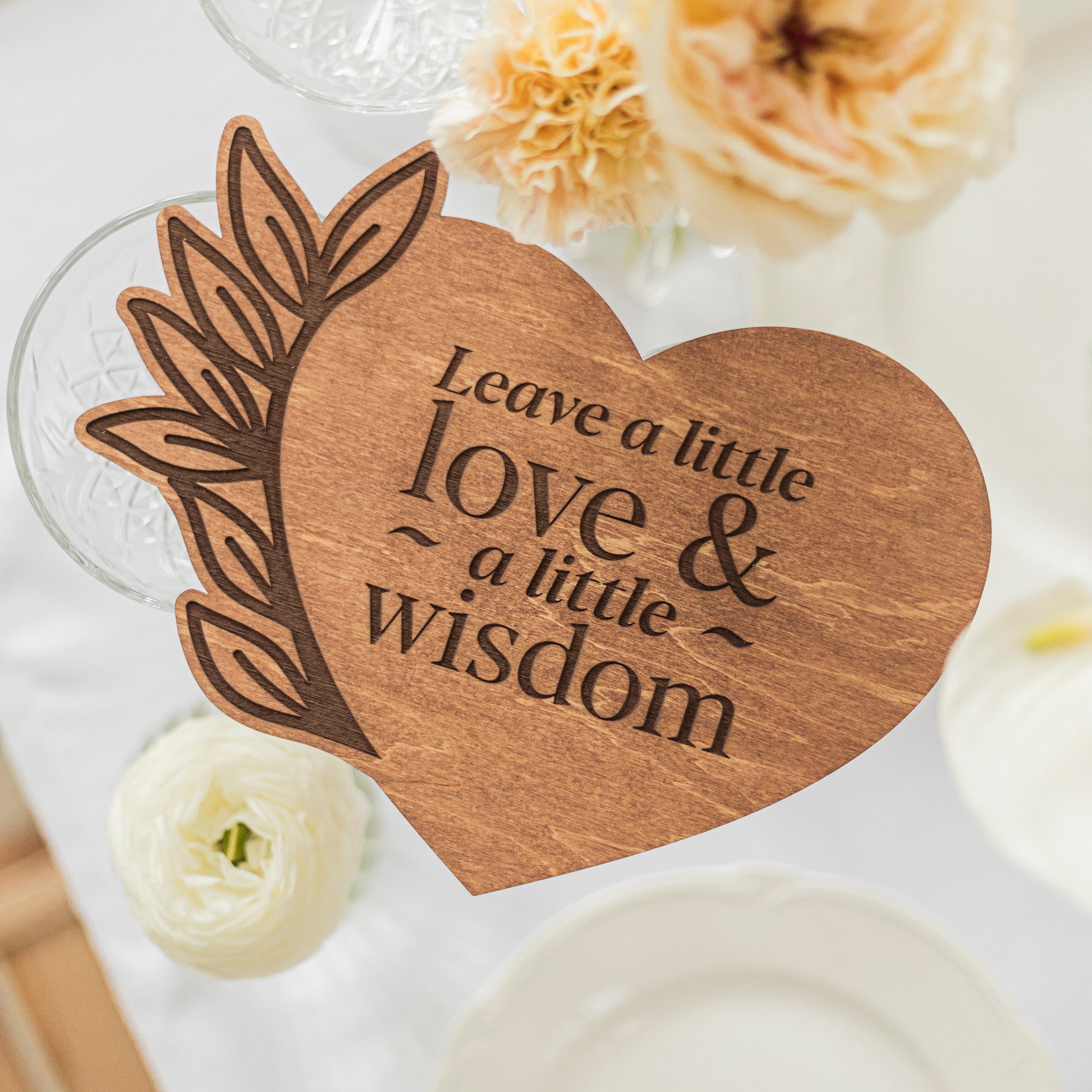 Wooden heart-shaped plaque with engraved text 'Leave a little love & a little wisdom' on a decorative background.