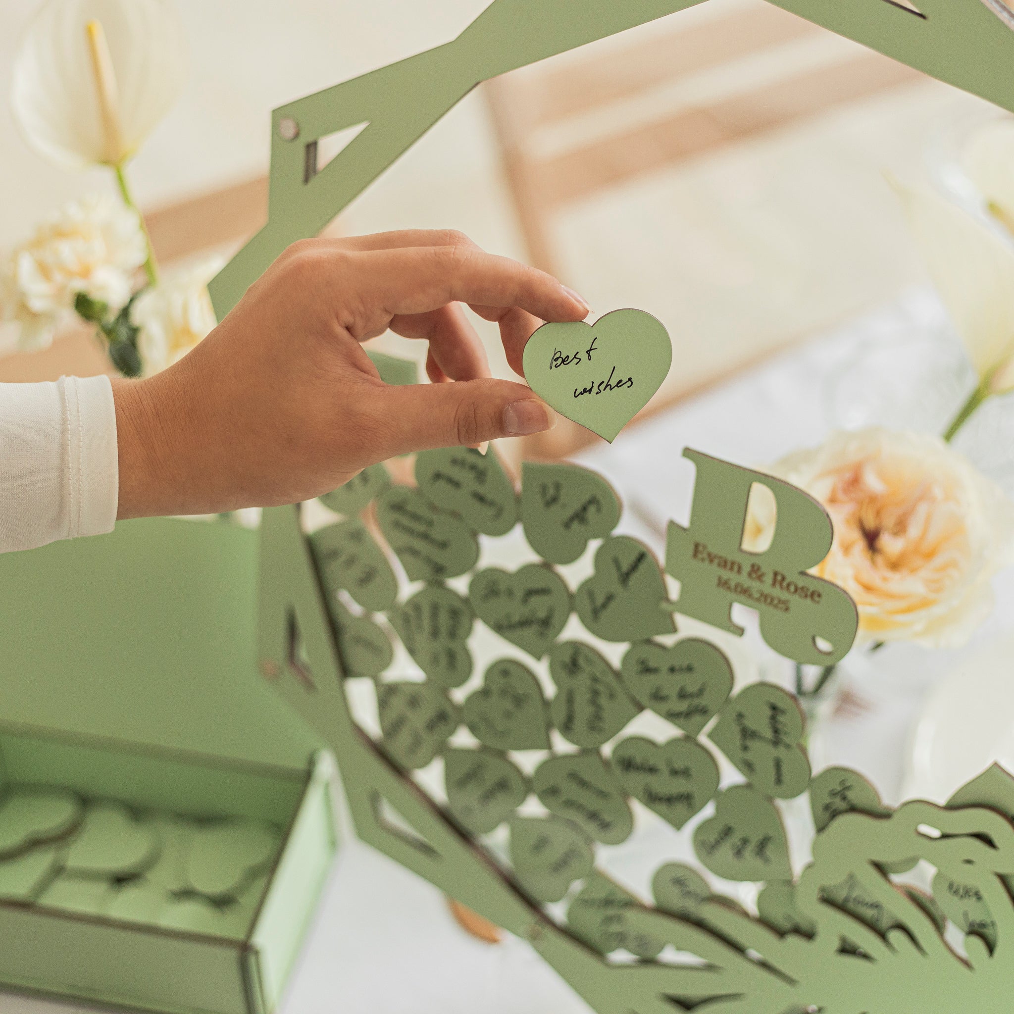 Hand holding a heart-shaped paper with 'Best wishes' text, surrounded by more heart-shaped papers in a decorative box.