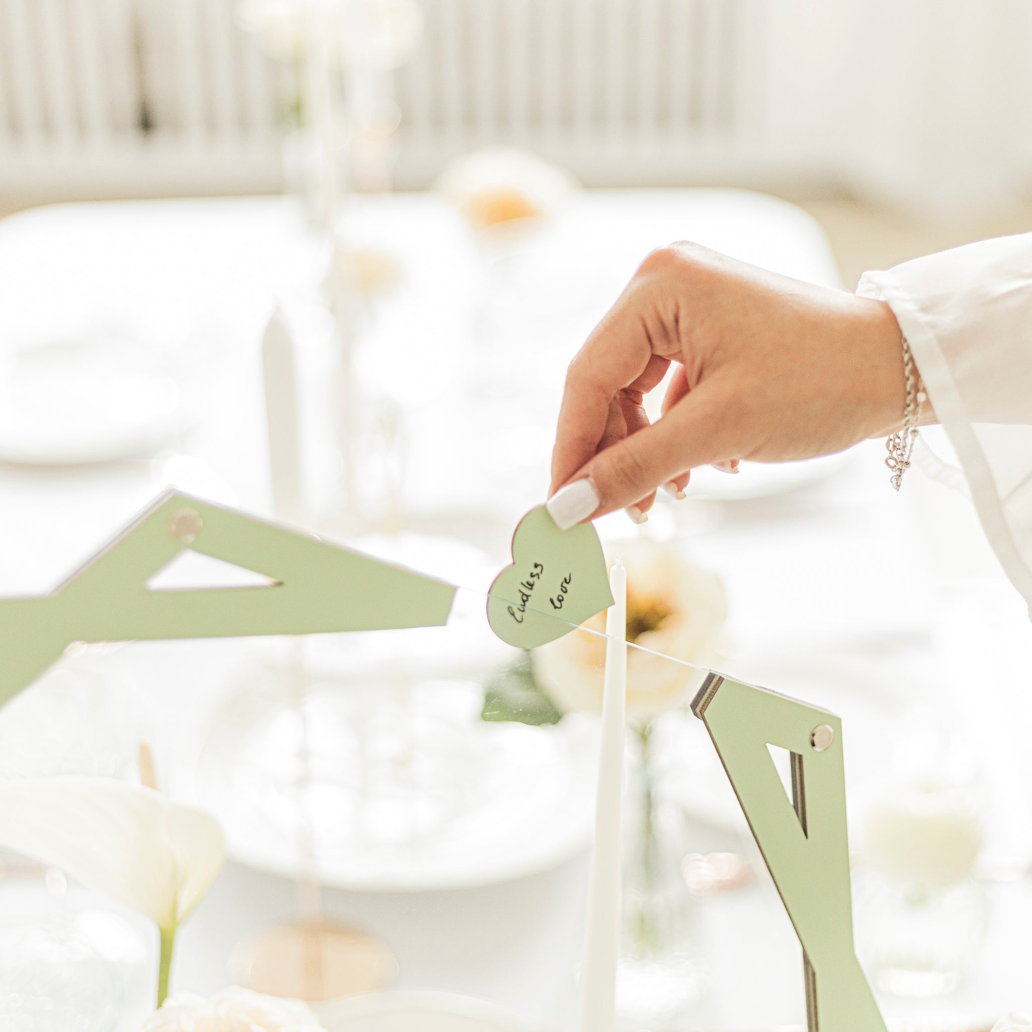Hand holding a heart-shaped card with 'Loves' on it, over a decorative stand with flowers in the background.