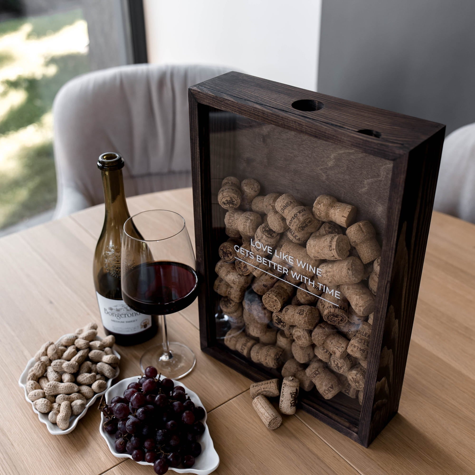 A dark wood wine cork display box with a glass front panel sits on a wooden table