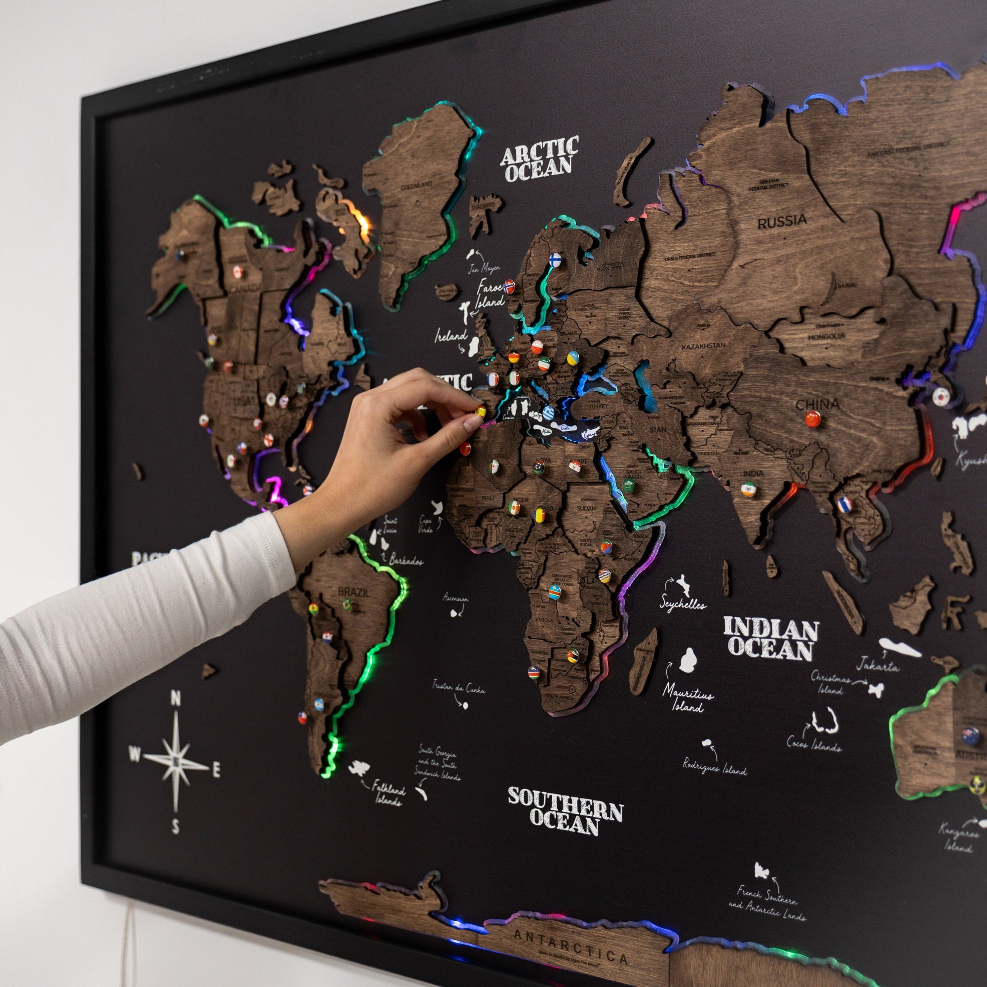 Wooden world map with colorful pins on a black background, hand pointing to a location.