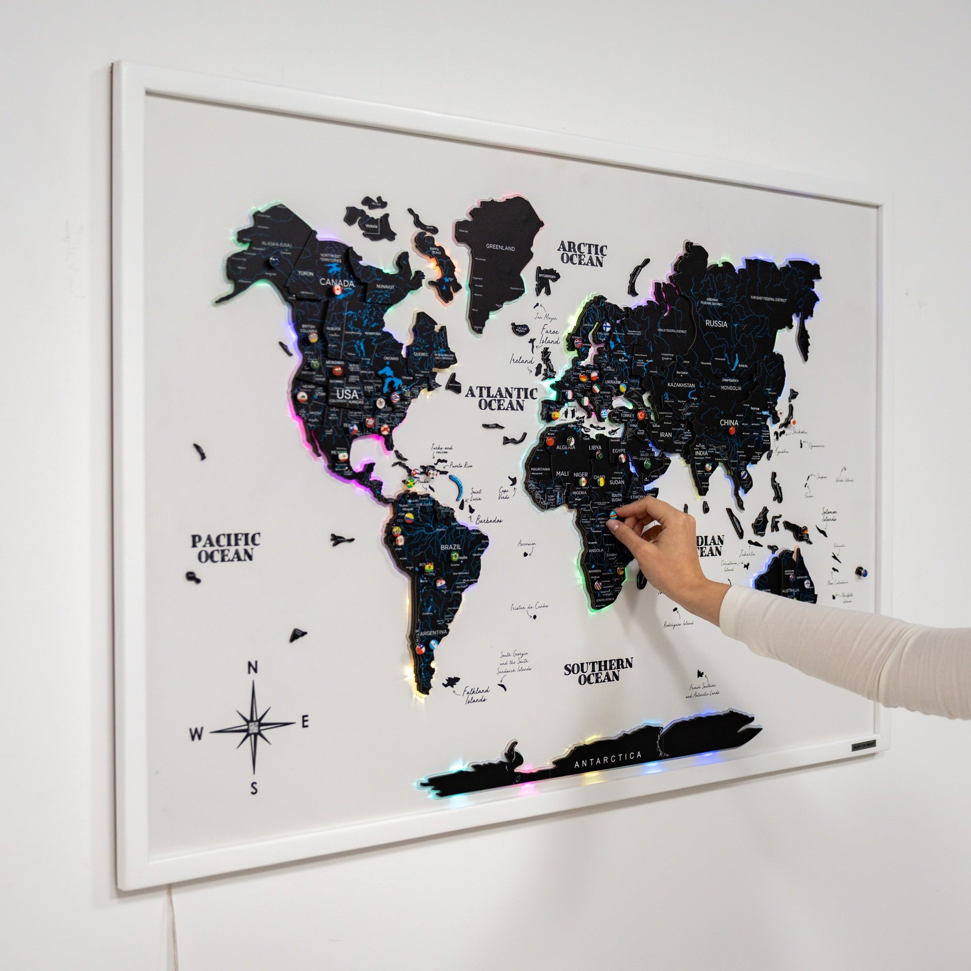 World map with colorful pins on a white wall, hand pointing at a location.