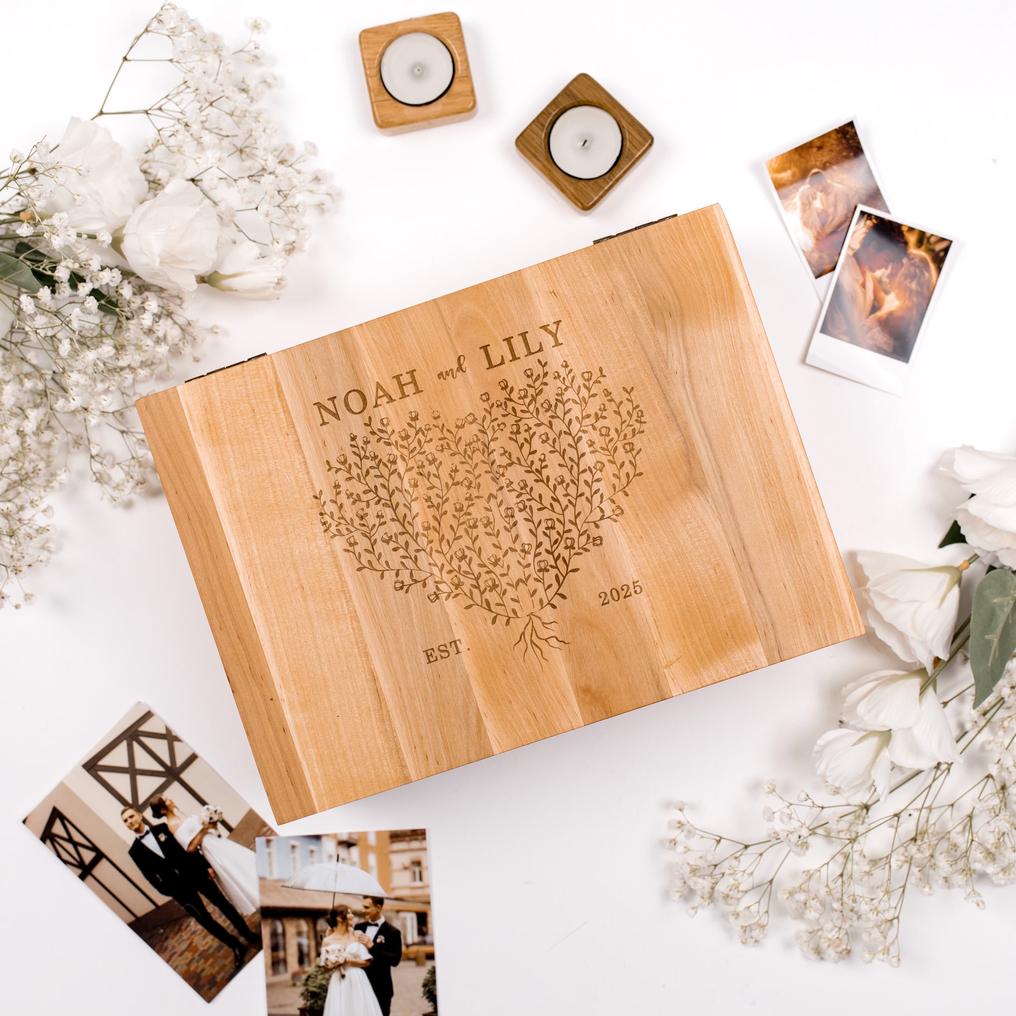 Wooden box with engraved design and text, surrounded by flowers and photos on a white surface