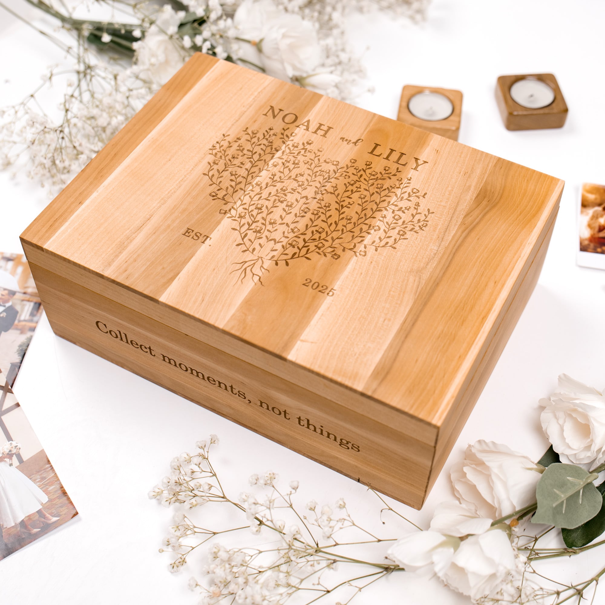 Wooden box with engraved text and design, surrounded by flowers and candles on a light background