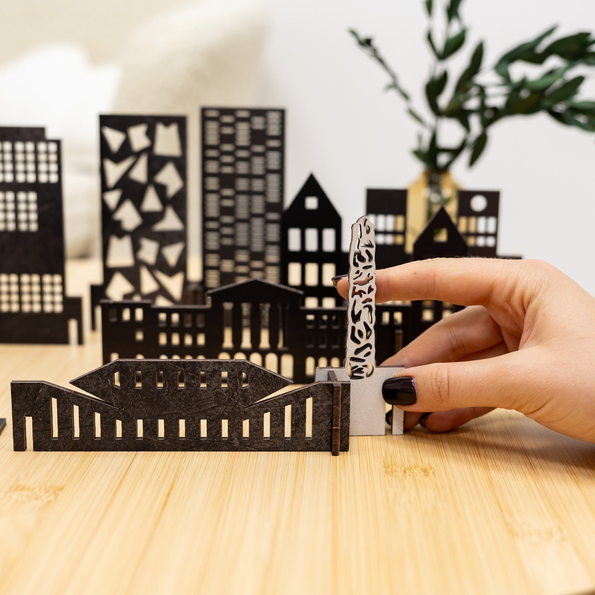 Hand holding a small metallic object in front of a black cut-out cityscape on a wooden surface.