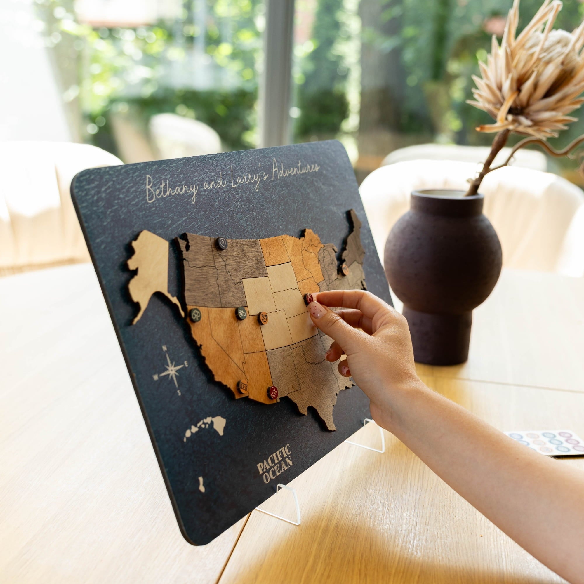 A wooden USA travel map puzzle board with a hand placing pins on states, displayed on a table in a bright indoor setting.