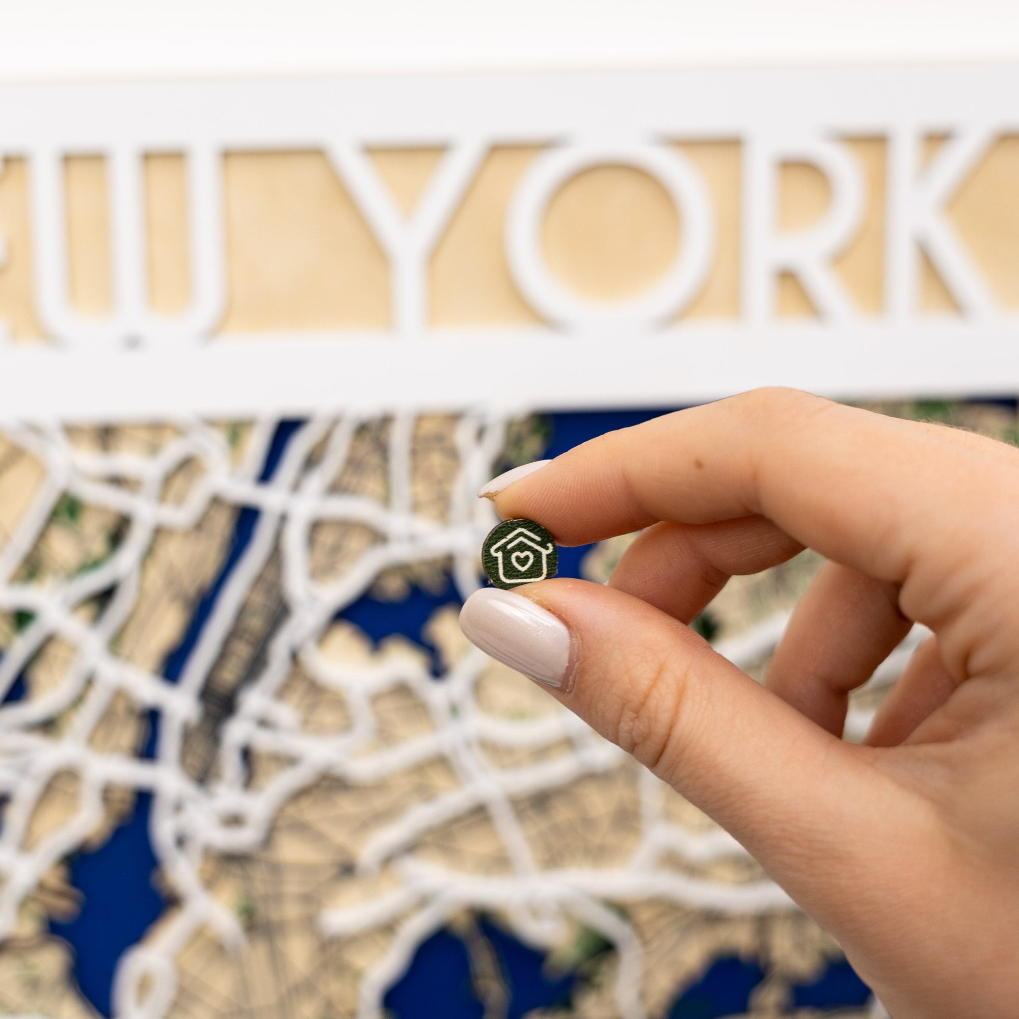 Hand holding a small black and gold house-shaped pin in front of a decorative wall with 'New York' text.