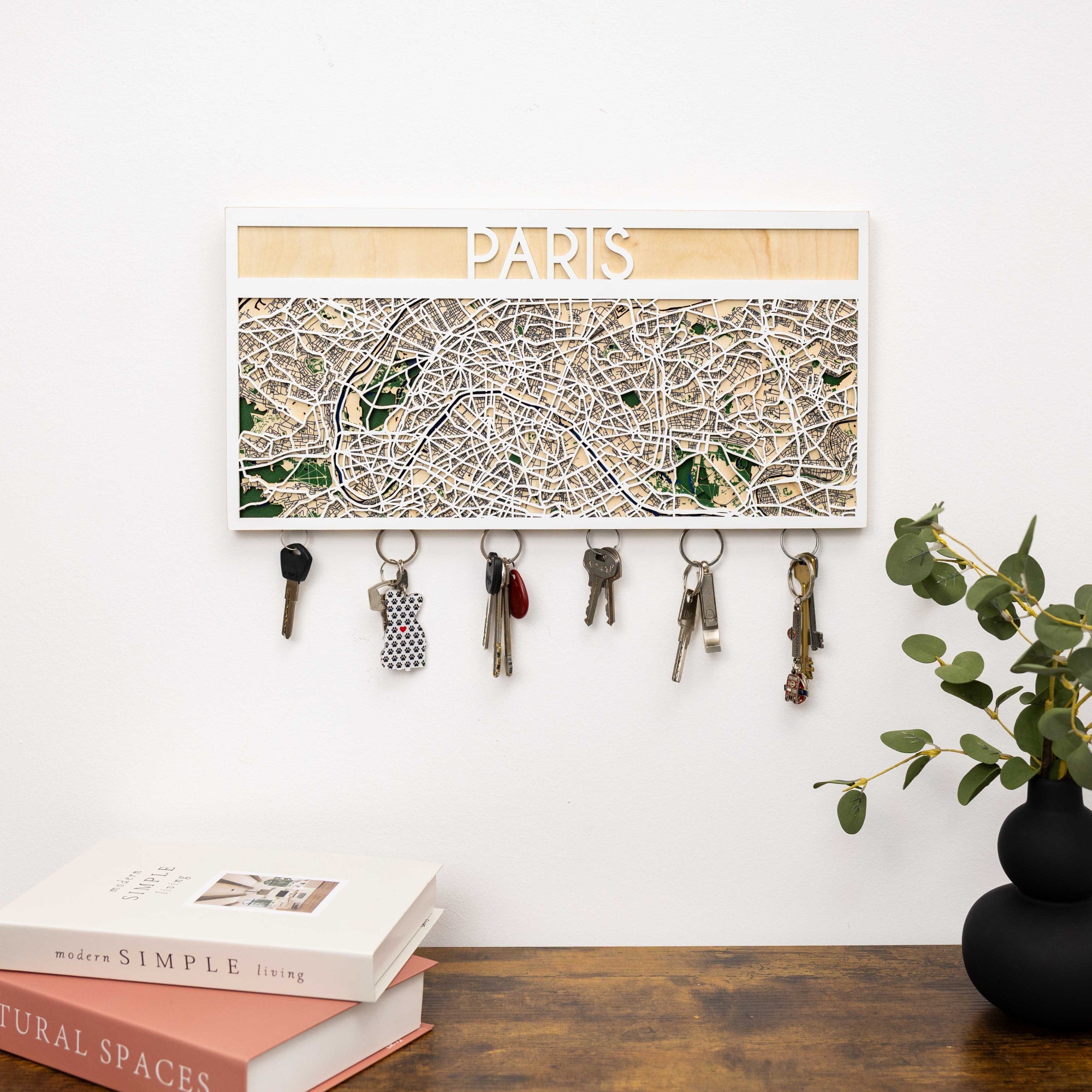 Key holder with Paris map design on a white wall above a wooden surface with books and a plant.