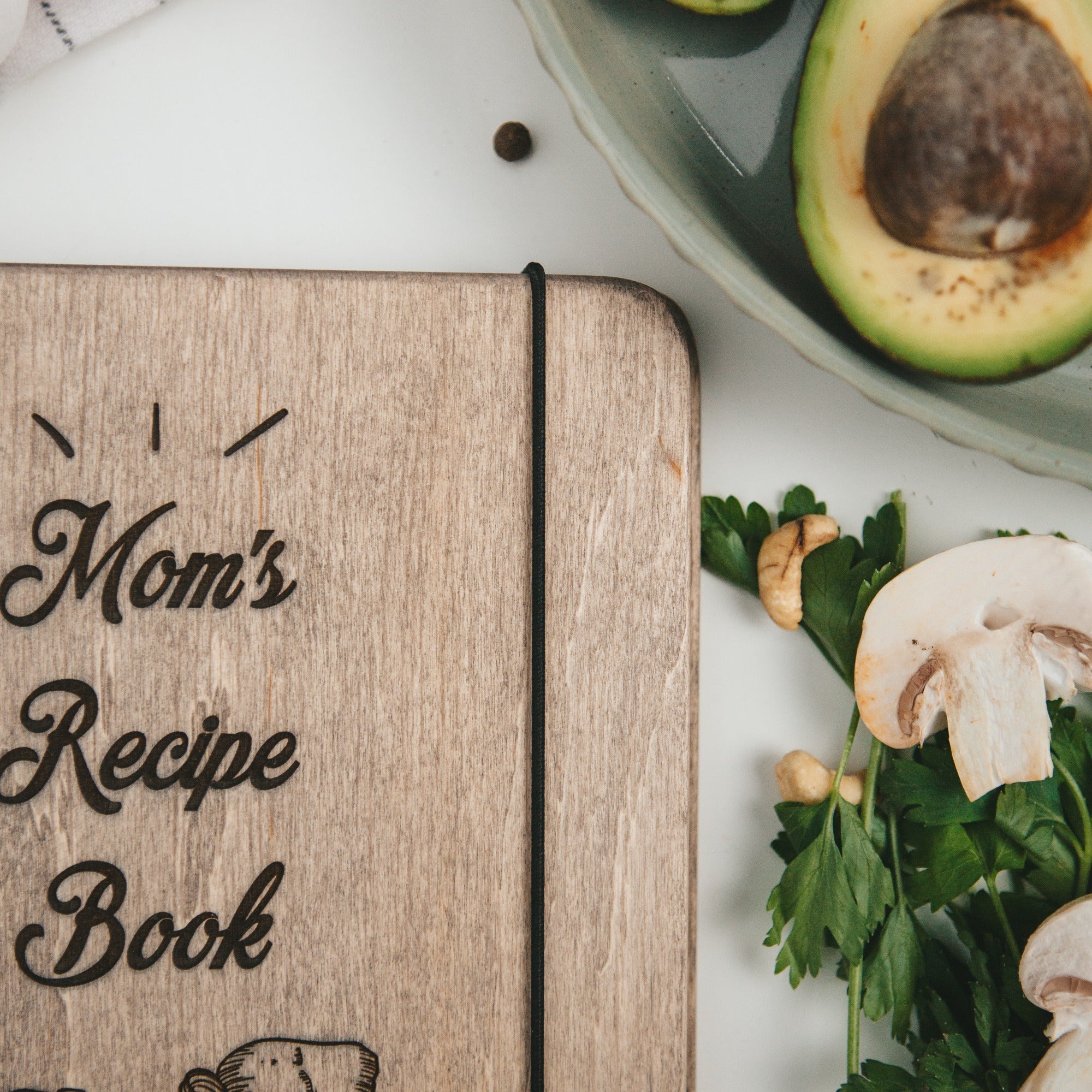 Wooden notebook titled 'Mom's Recipe Book' with ingredients like mushrooms and parsley on a white surface.