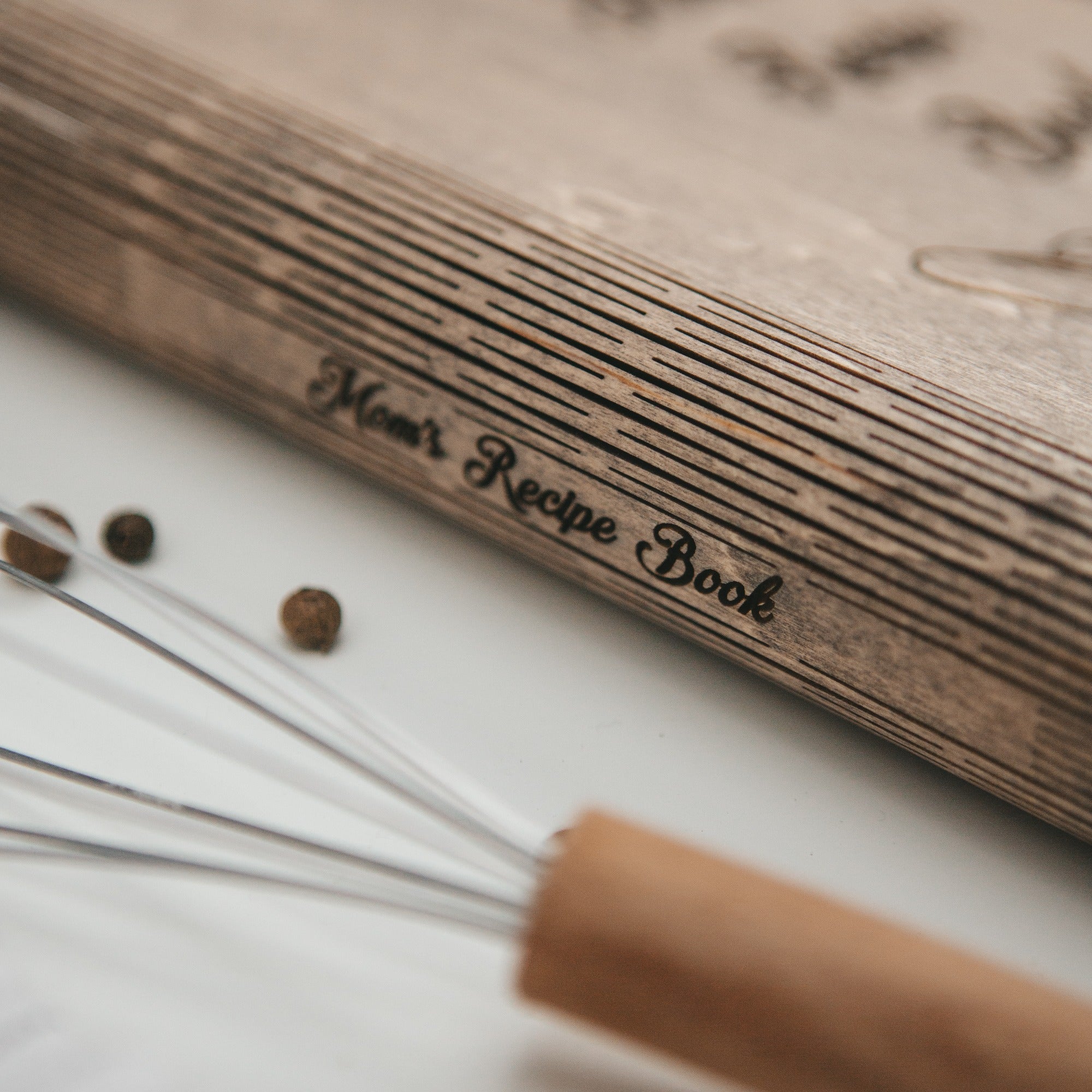 Wooden recipe book titled 'Mom's Recipe Book' with a whisk and spices on a white surface.