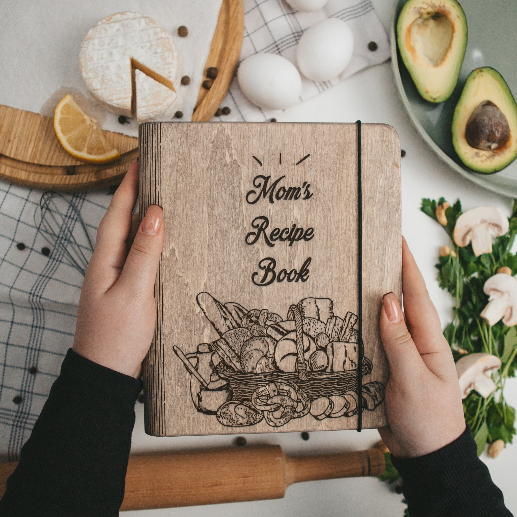 Wooden recipe book titled 'Mom's Recipe Book' held by hands with kitchen items in the background.