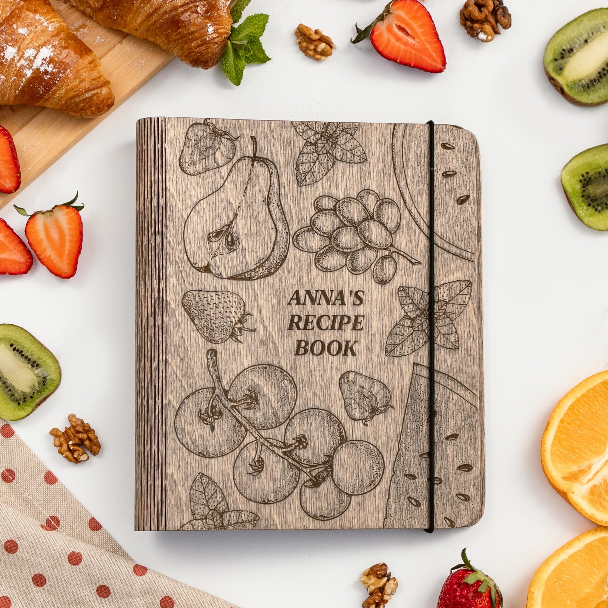 Wooden recipe book titled 'Anna's Recipe Book' surrounded by fruits and pastries on a white surface.