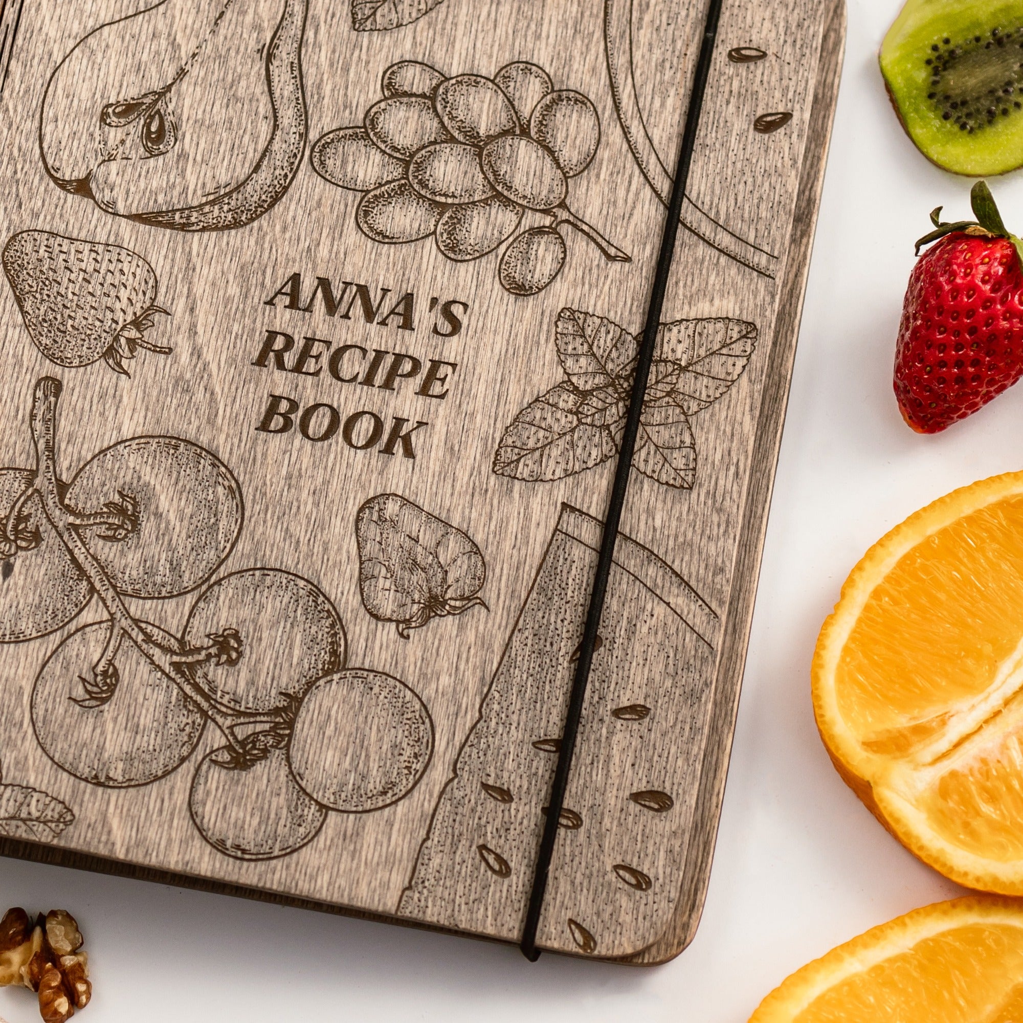 Wooden recipe book with fruit illustrations and text 'Anna's Recipe Book' on a white background with fruits.