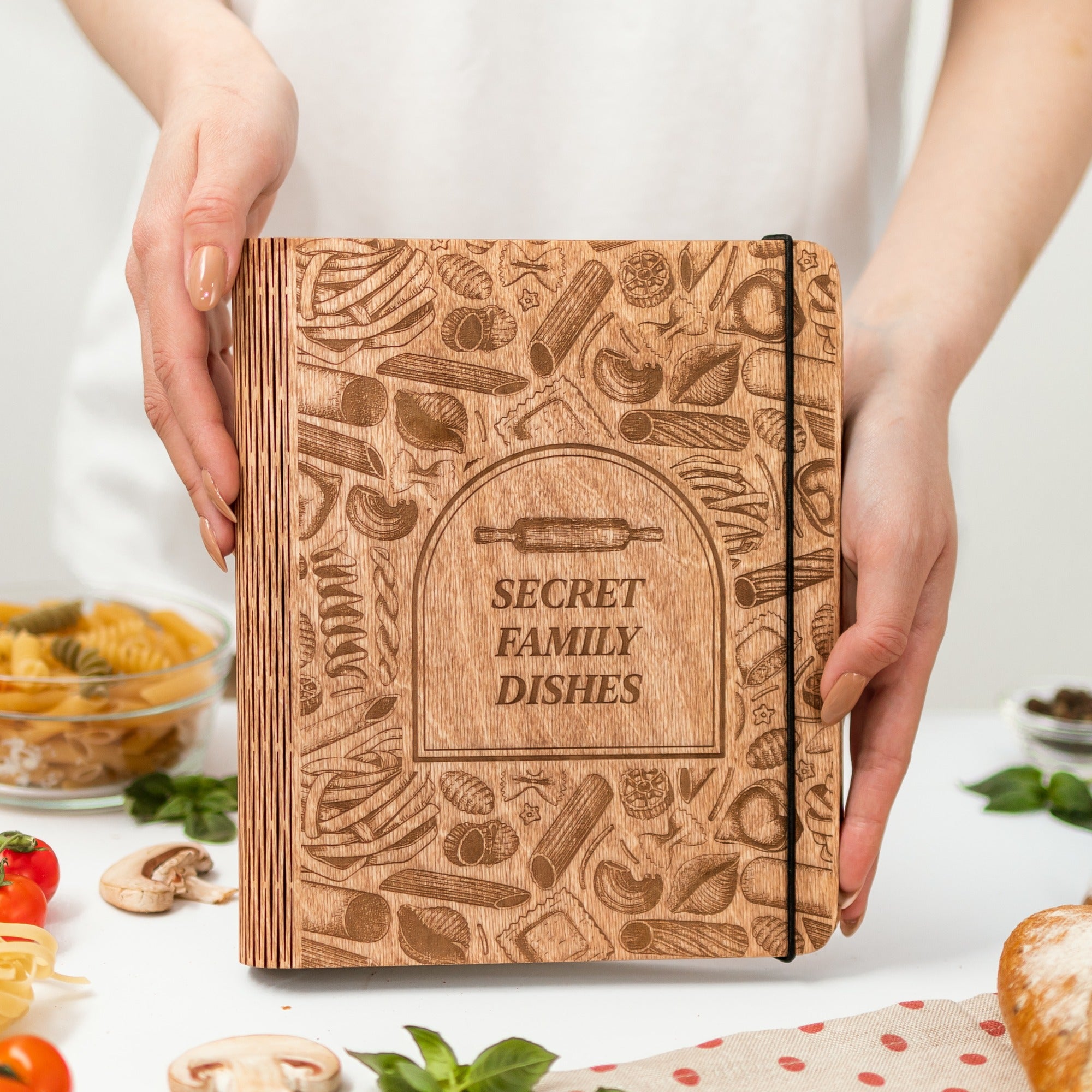 Wooden notebook with 'Secret Family Dishes' text held by hands, surrounded by kitchen items.
