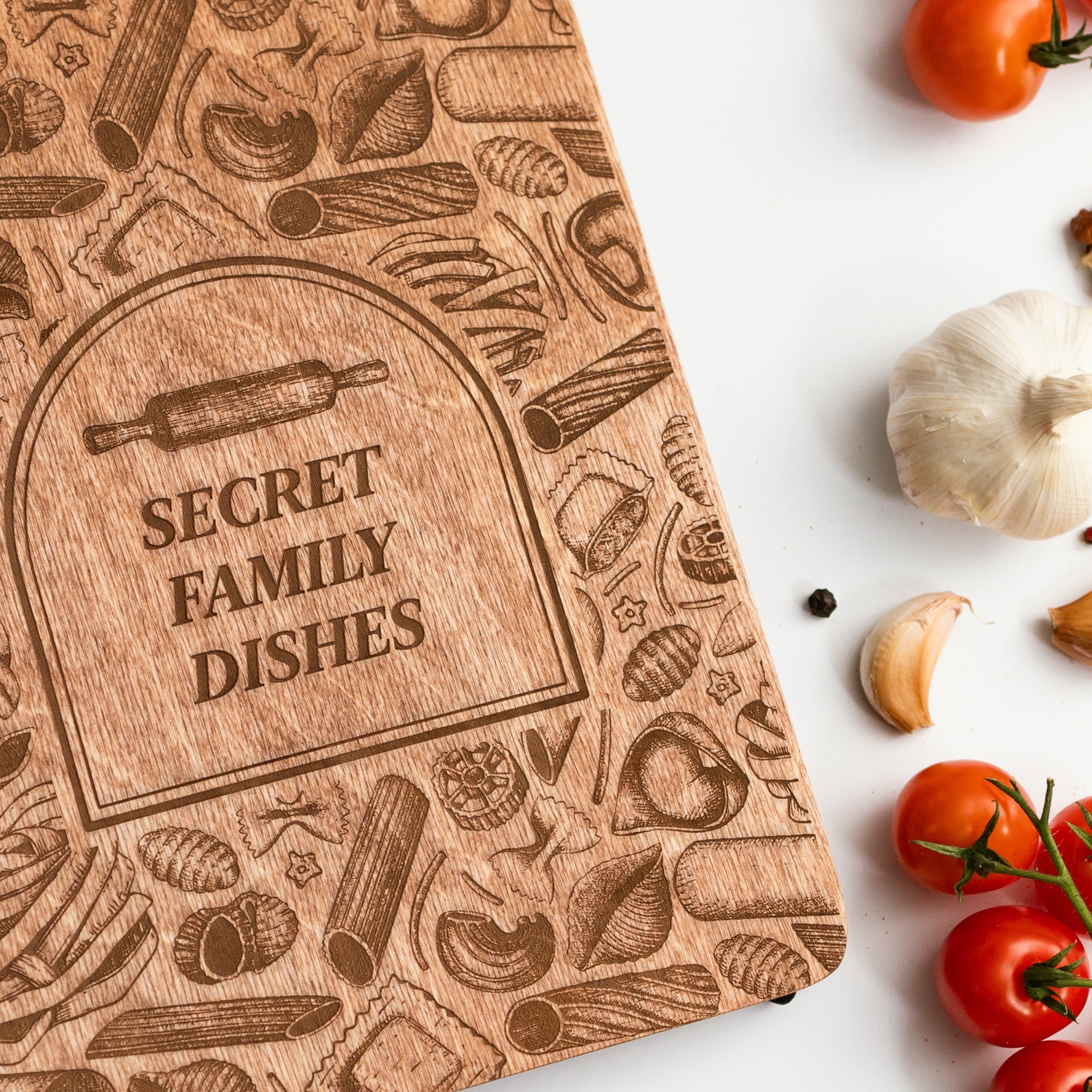 Wooden cutting board with engraved patterns and 'Secret Family Dishes' text, surrounded by tomatoes and garlic on a white surface.
