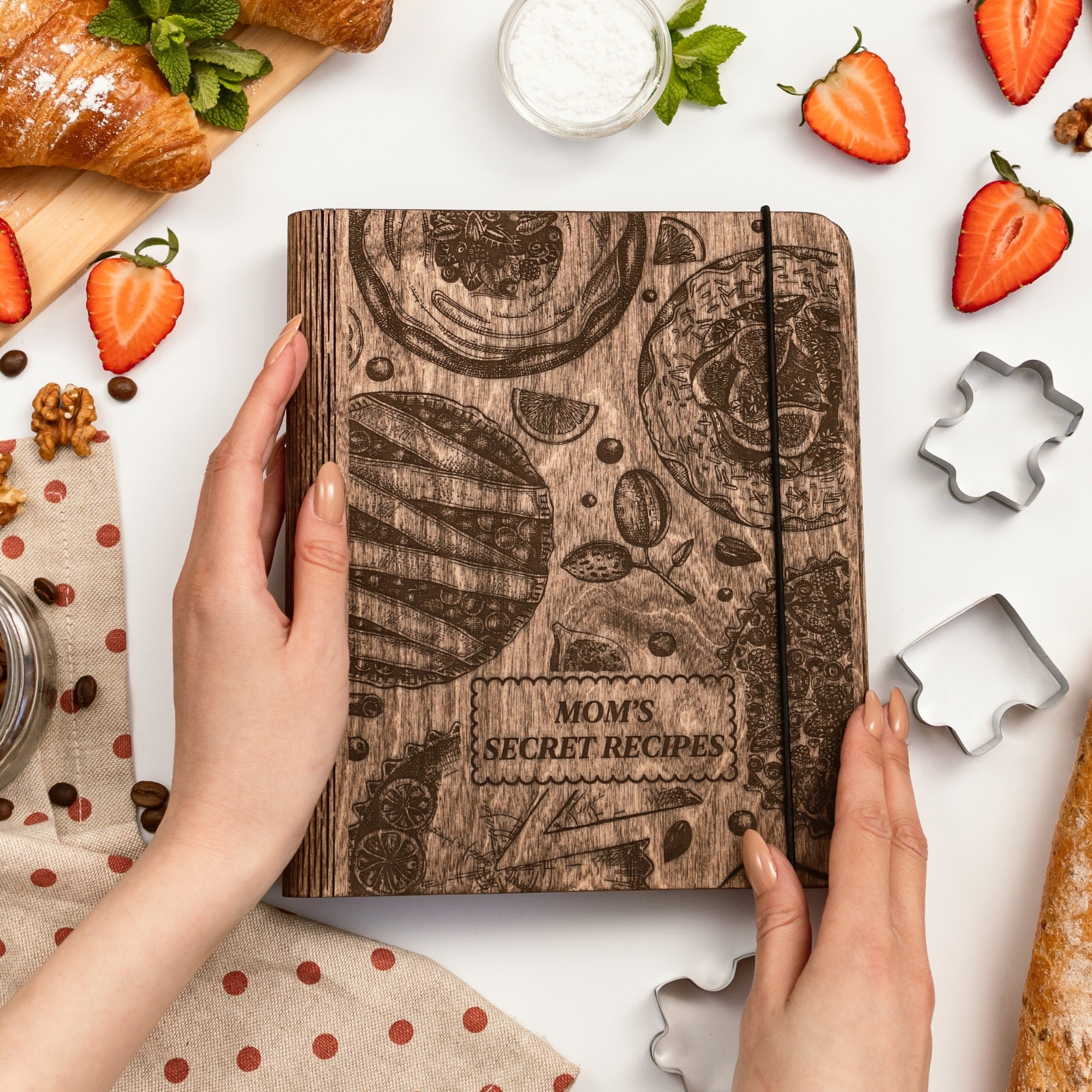 Cookbook titled 'Mom's Secret Recipes' held by a hand with kitchen items around on a white background