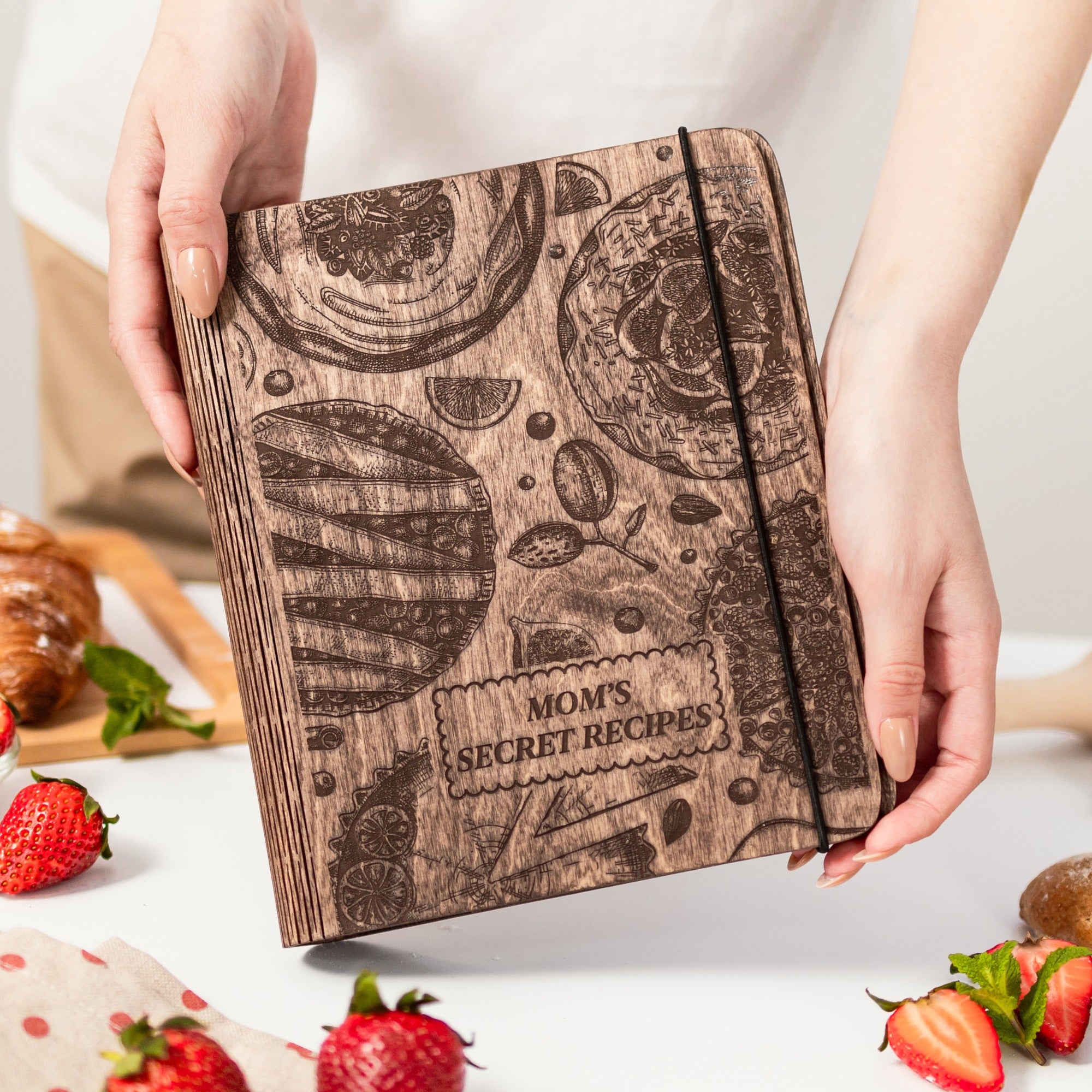 Wooden recipe book titled 'Mom's Secret Recipes' held by hands with strawberries and pastries in the background.
