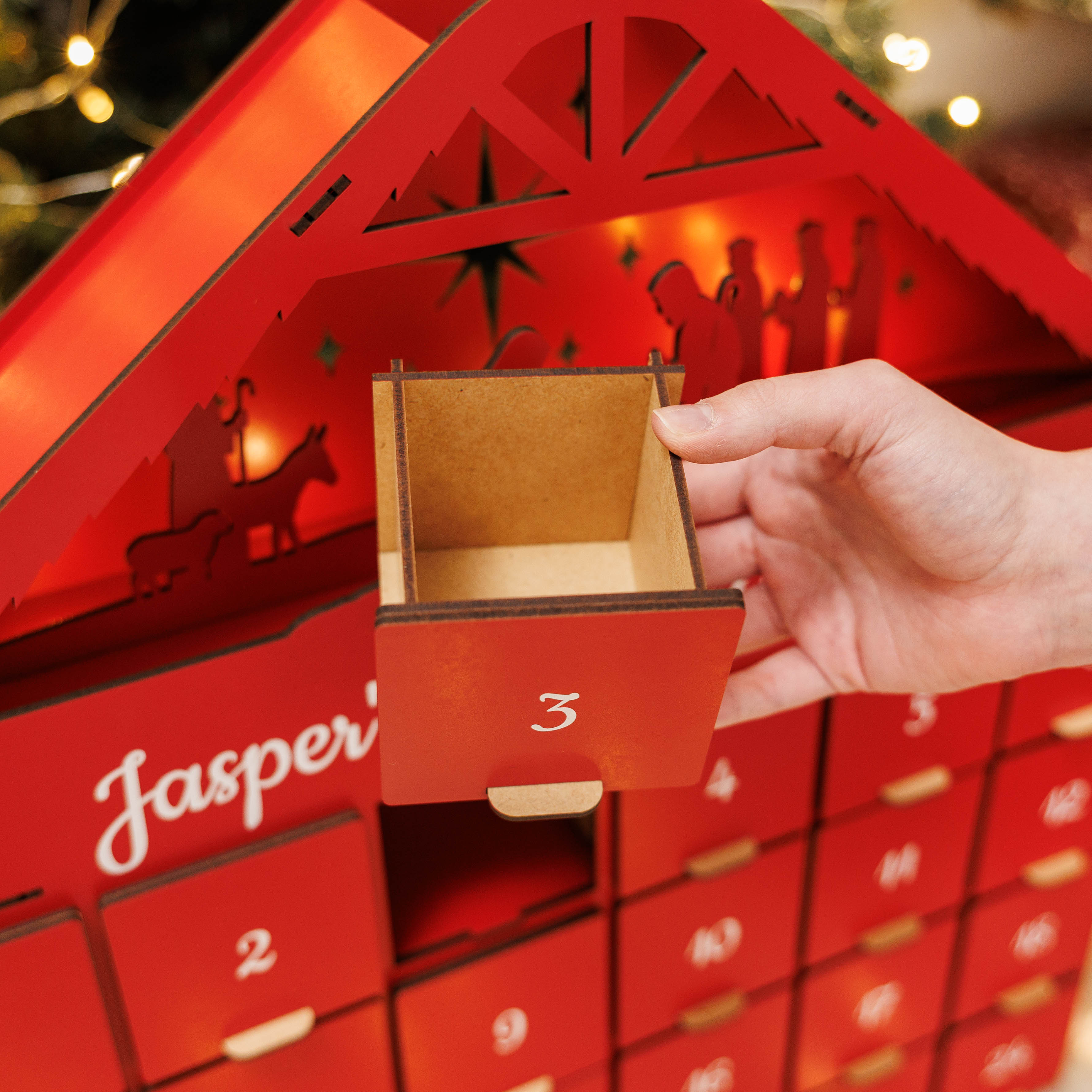 Red wooden advent calendar with a hand opening a door, Christmas lights in the background