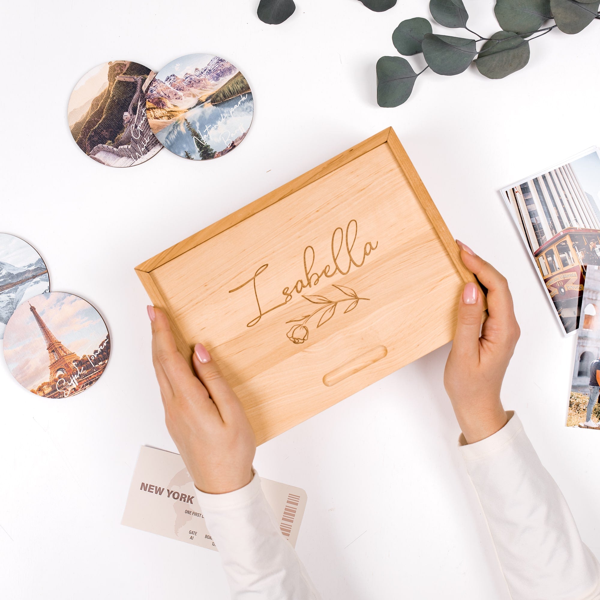 Wooden box with 'Isabella' engraving held by hands on a white surface with scenic photos and greenery.