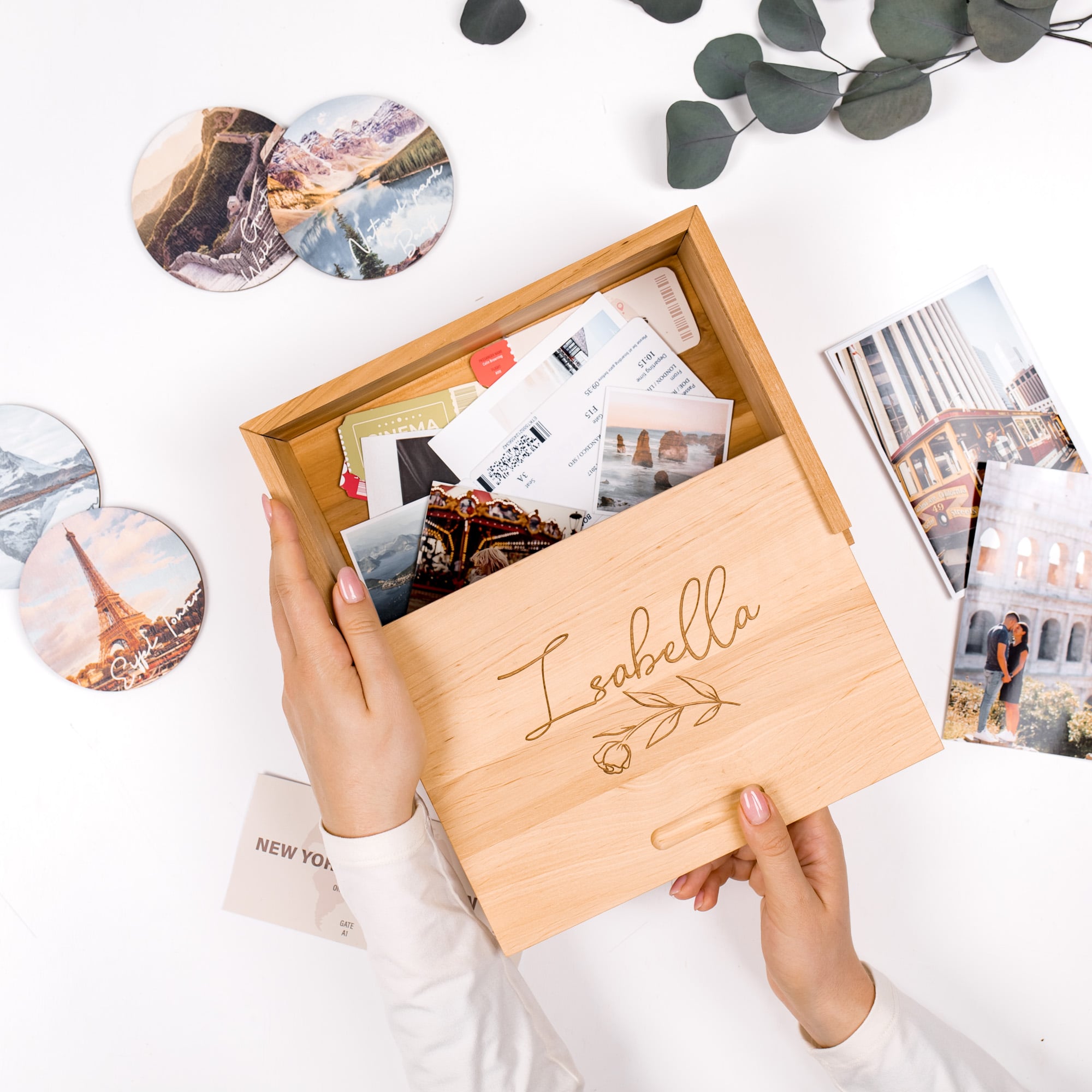 Wooden box with 'Isabella' engraving, held by a person, with travel-themed photos and coasters on a white surface.
