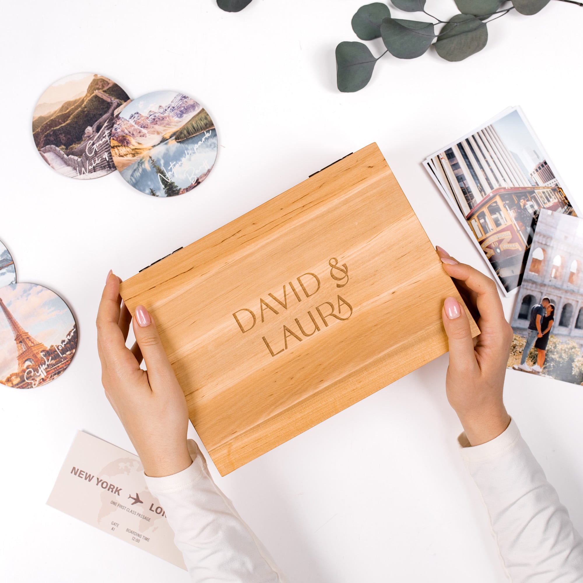 Wooden box with 'DAVID & LAURA' engraving held by hands on a white surface with travel-themed items.